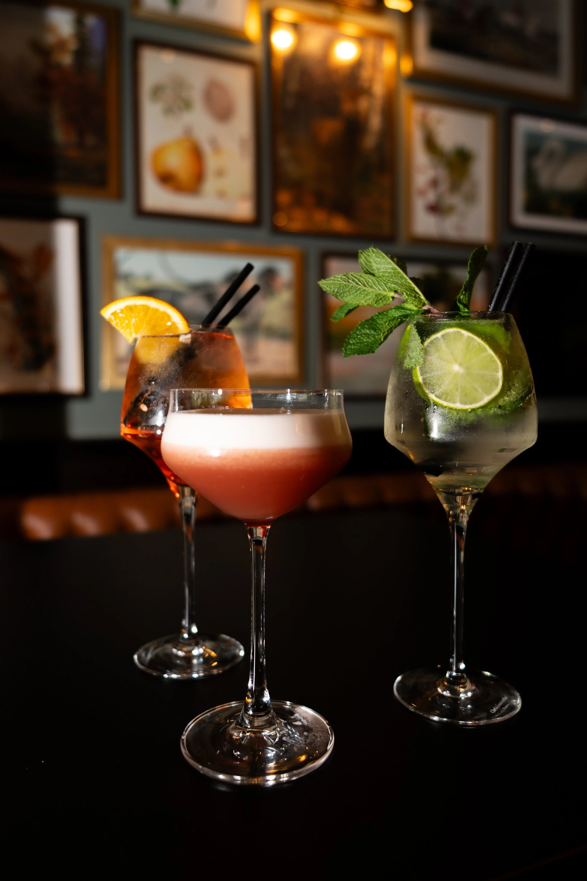 Three cocktail drinks with garnishes on a black table against a background of framed pictures.
