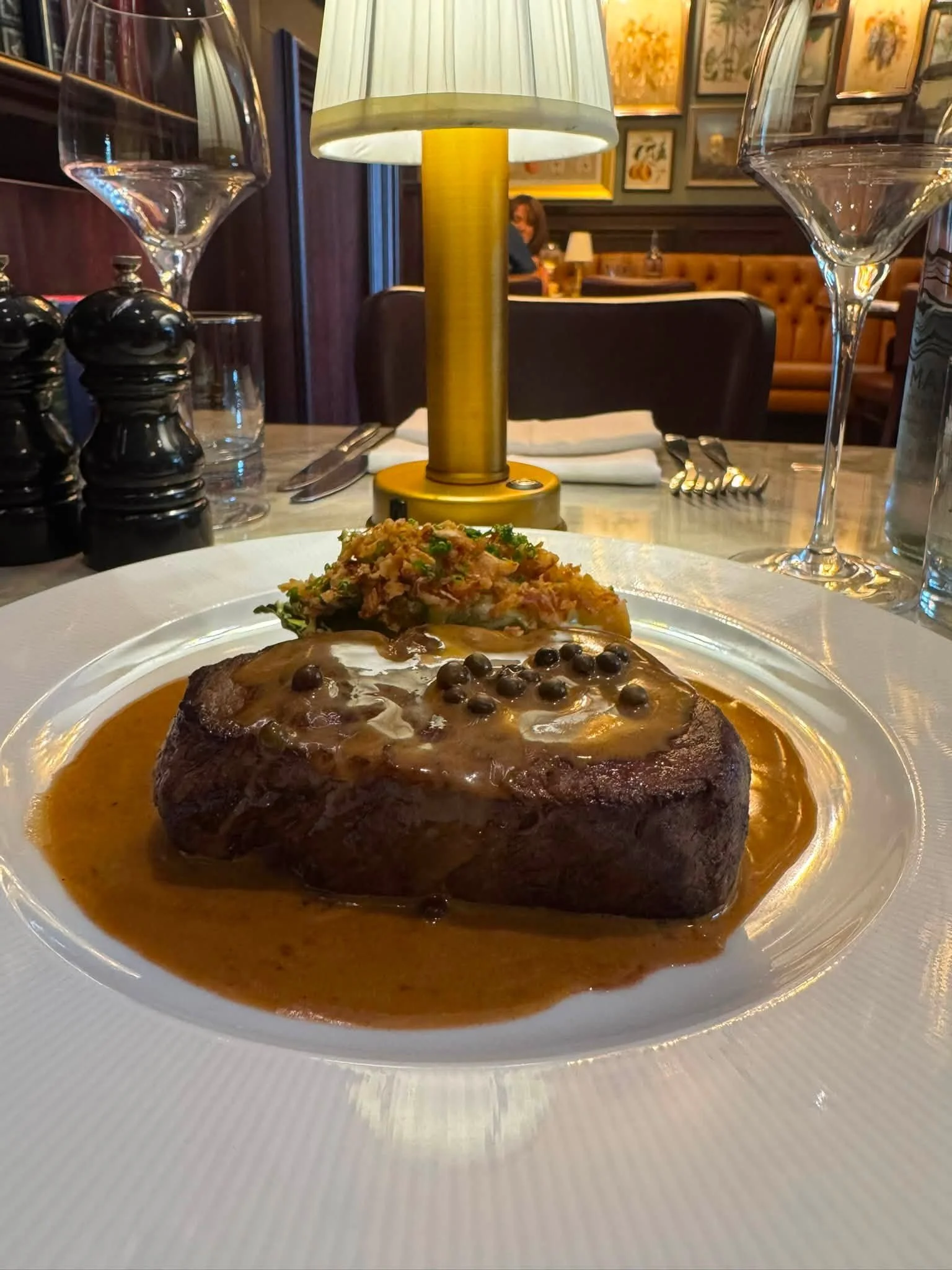 A plate of steak with mushroom sauce, topped with crispy onions and chopped herbs, served with a side of vegetables. The background shows a restaurant setting with a table lamp and framed artwork on the wall.