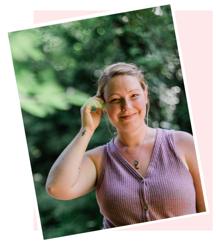 A woman smiling outdoors, adjusting her hair, with trees and greenery in the background.
