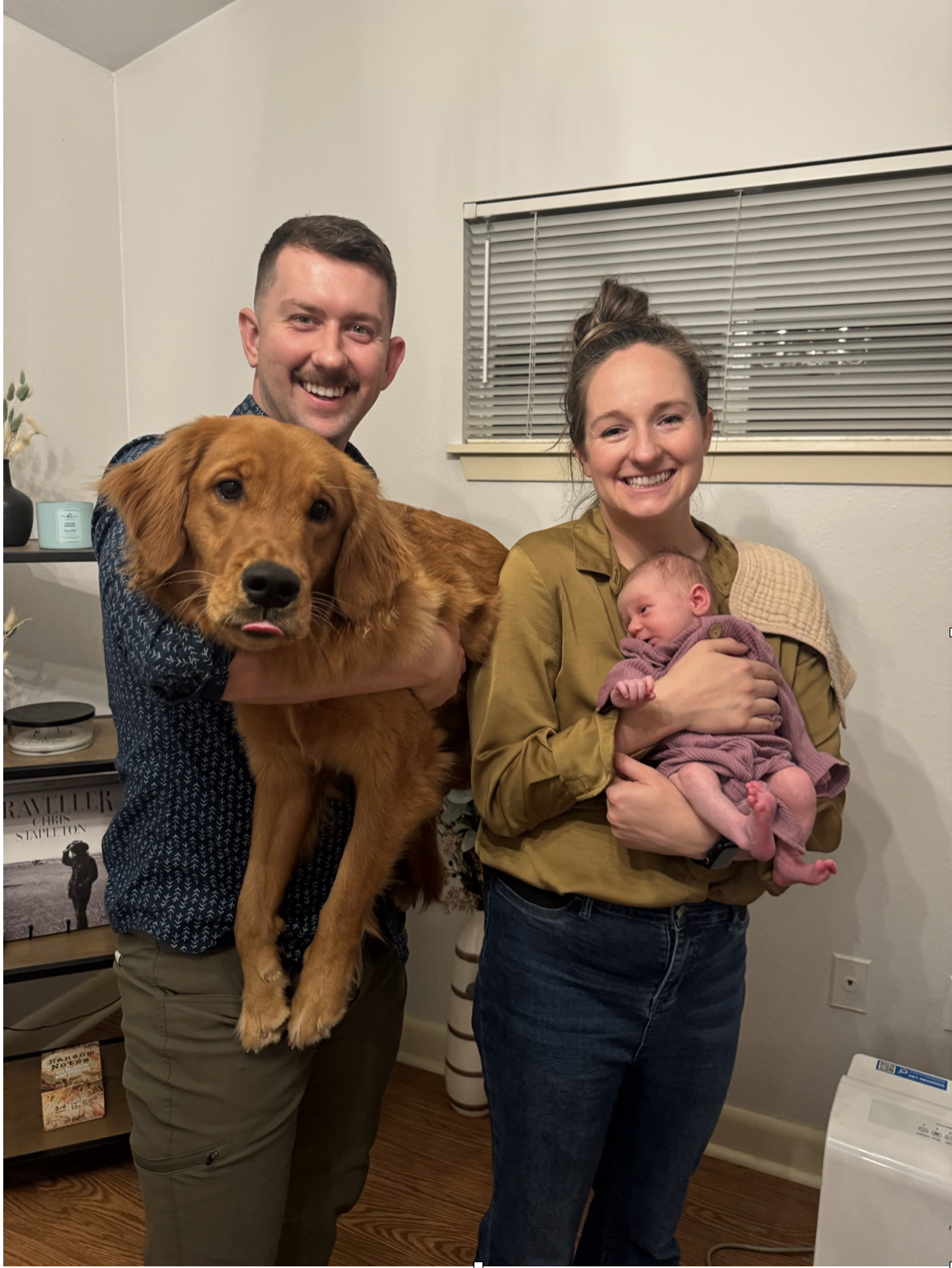 A happy man holding a golden retriever and a smiling woman holding a newborn baby in a cozy home setting.