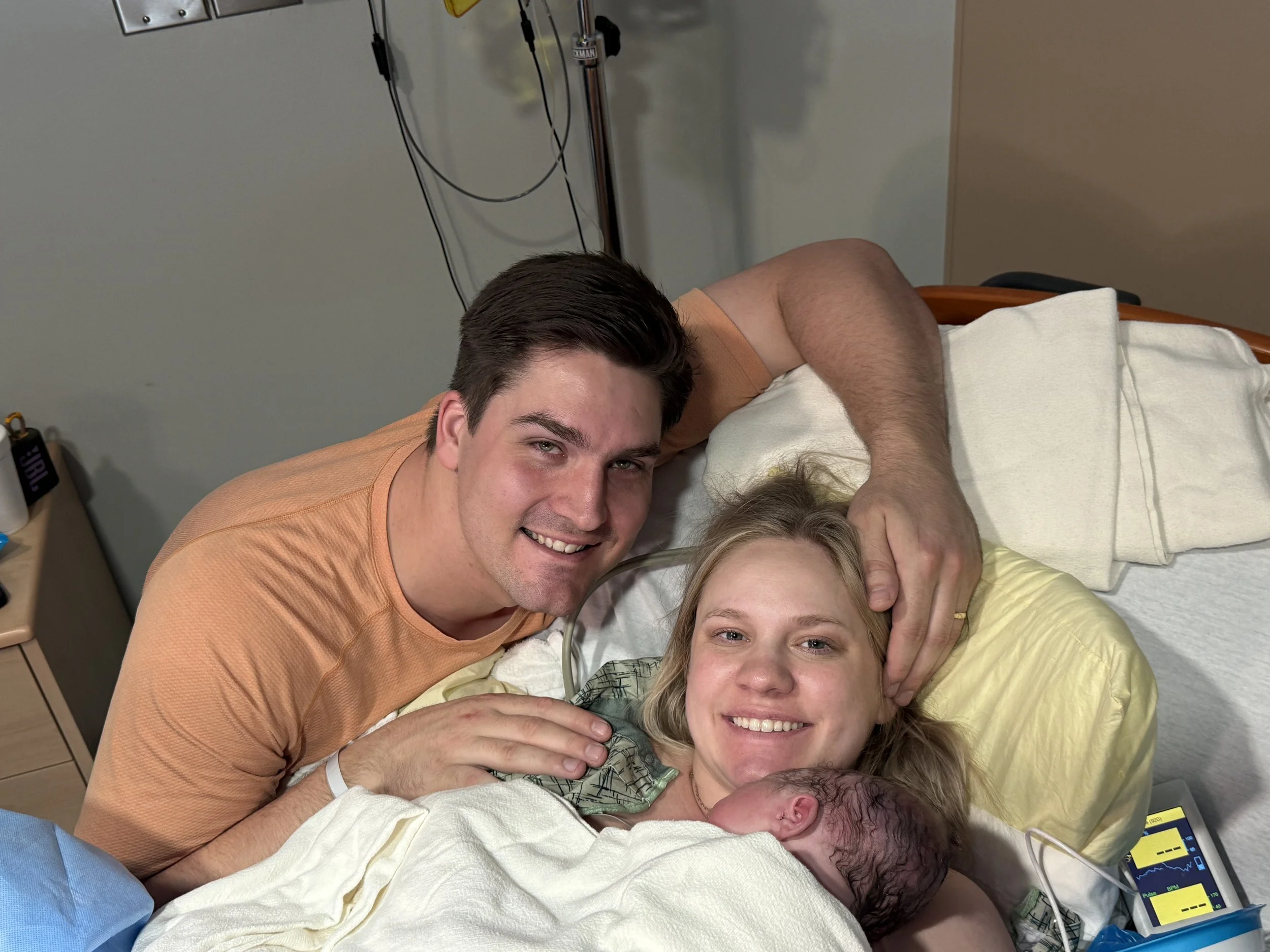 New mother lying in hospital bed holding newborn baby, with father leaning over and smiling.