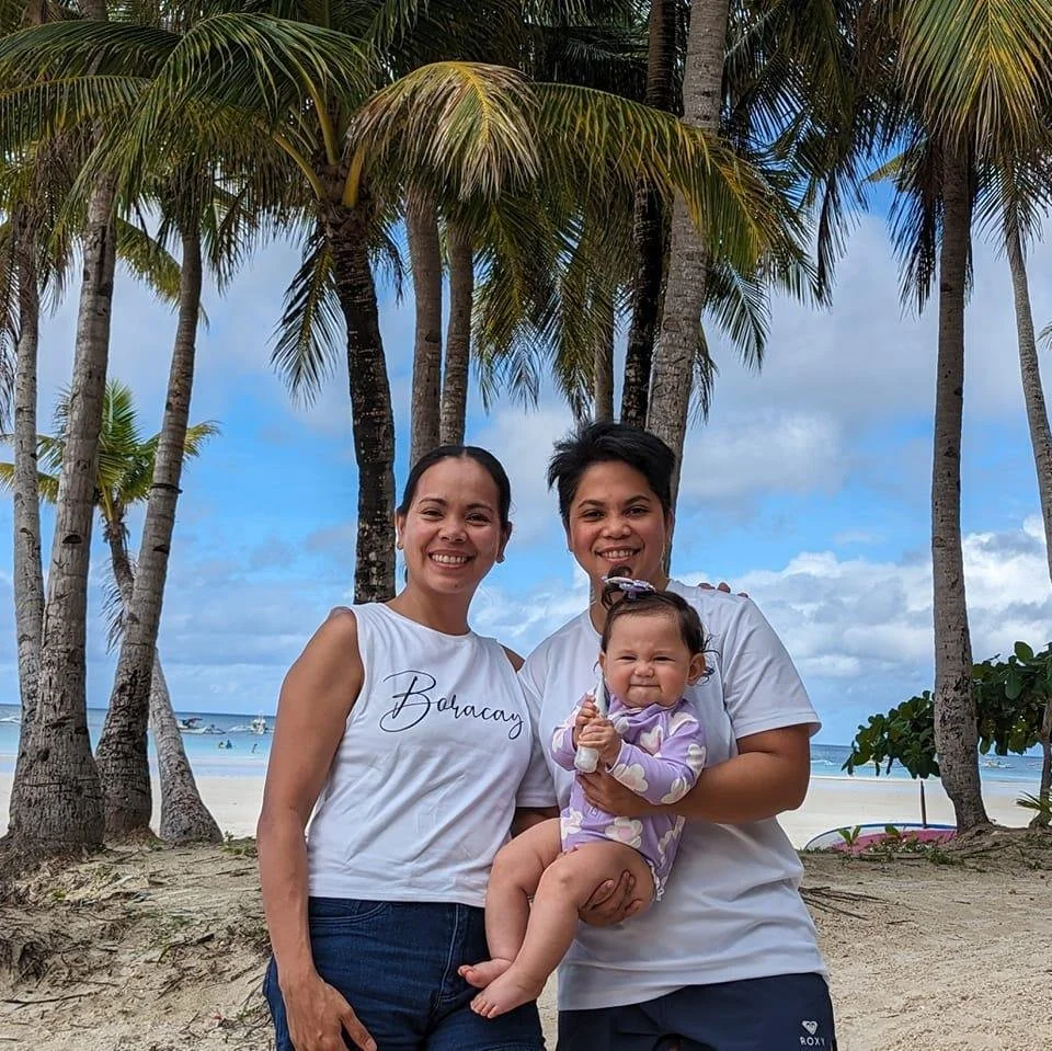 Three people smiling on a sandy beach with palm trees and blue sky in the background, two women and a young girl, one woman wearing a white sleeveless shirt and the other a white t-shirt, the girl wearing a purple and white outfit.