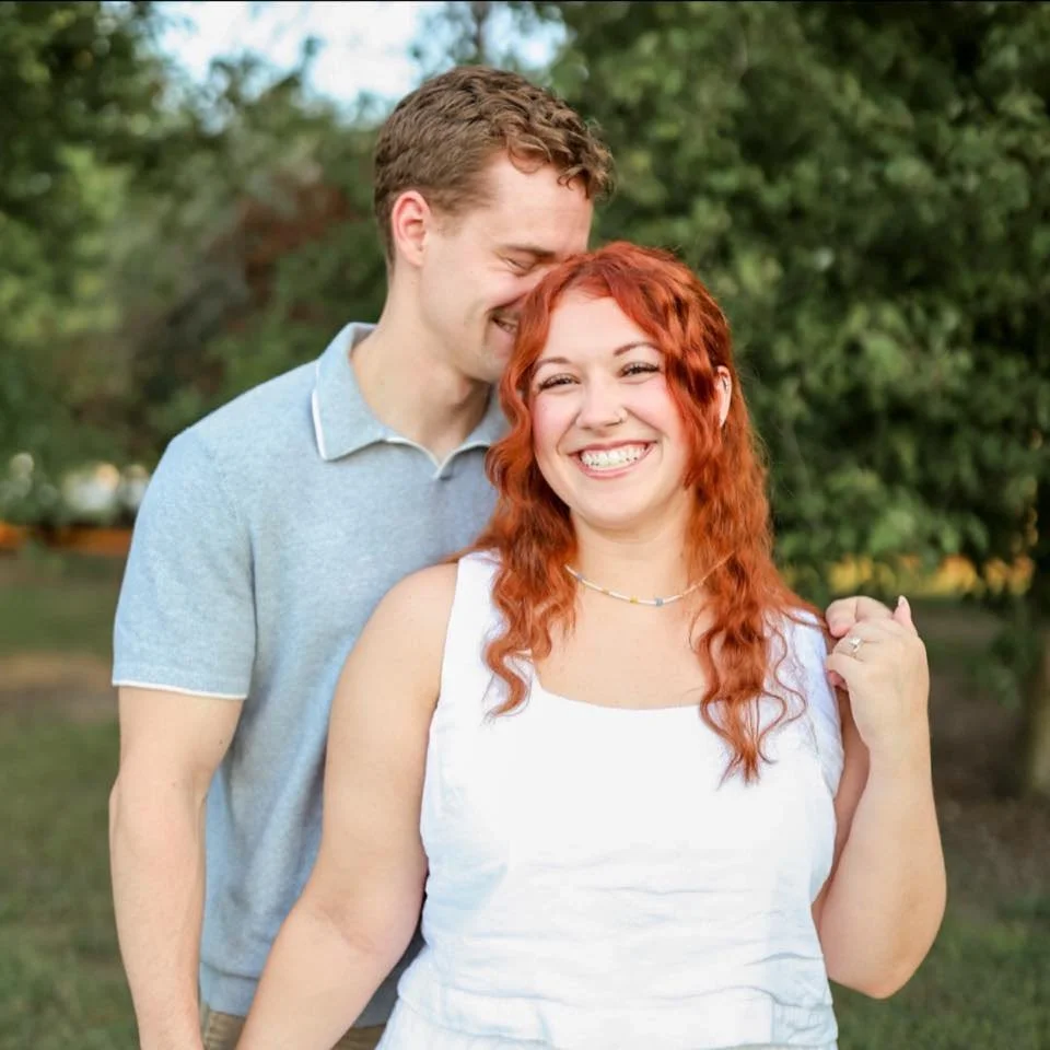 A smiling man and woman standing outdoors with trees in the background, the man leaning his head on the woman's forehead.