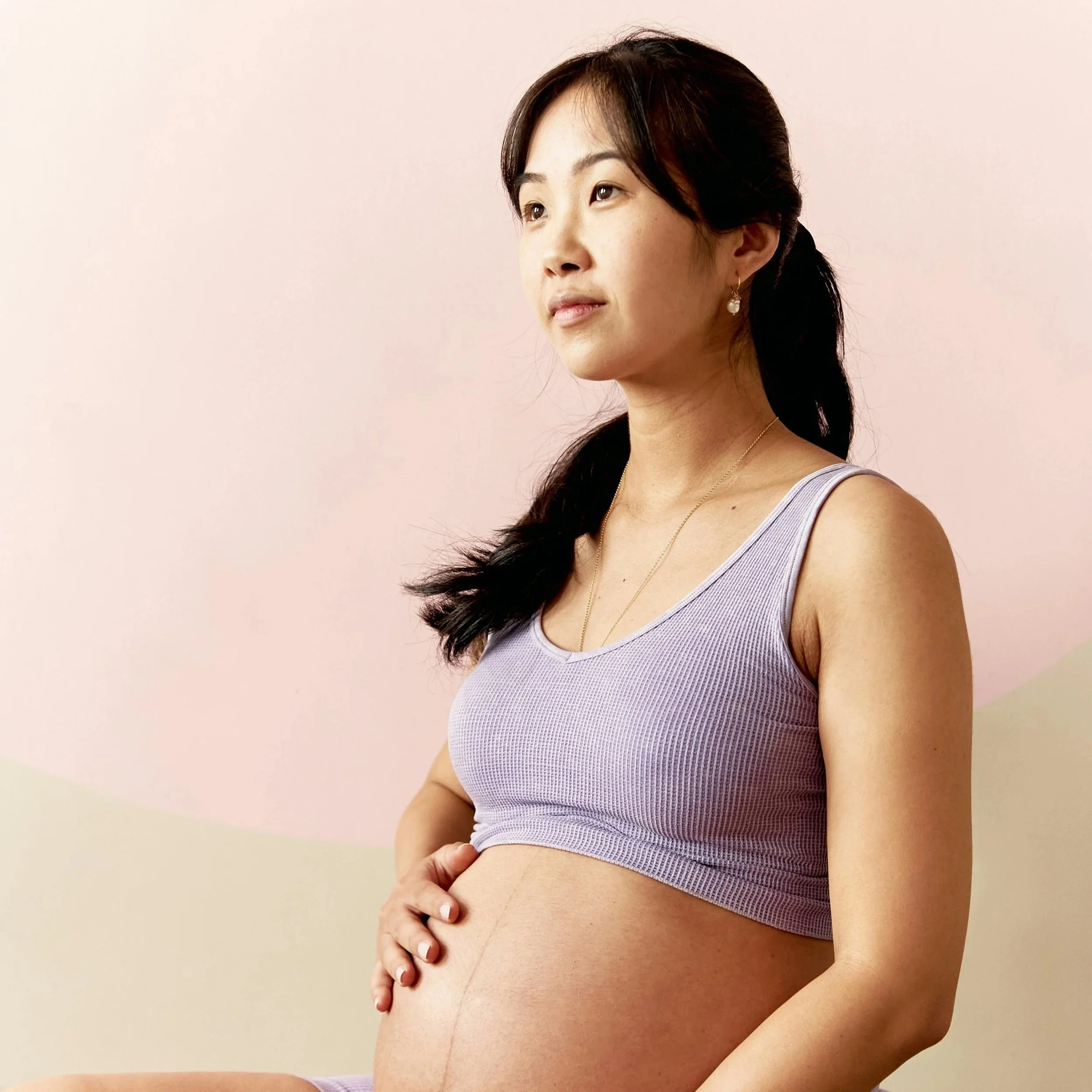 A pregnant woman wearing a lavender tank top and pearl earrings, posing with her hand on her belly, against a soft pink background.