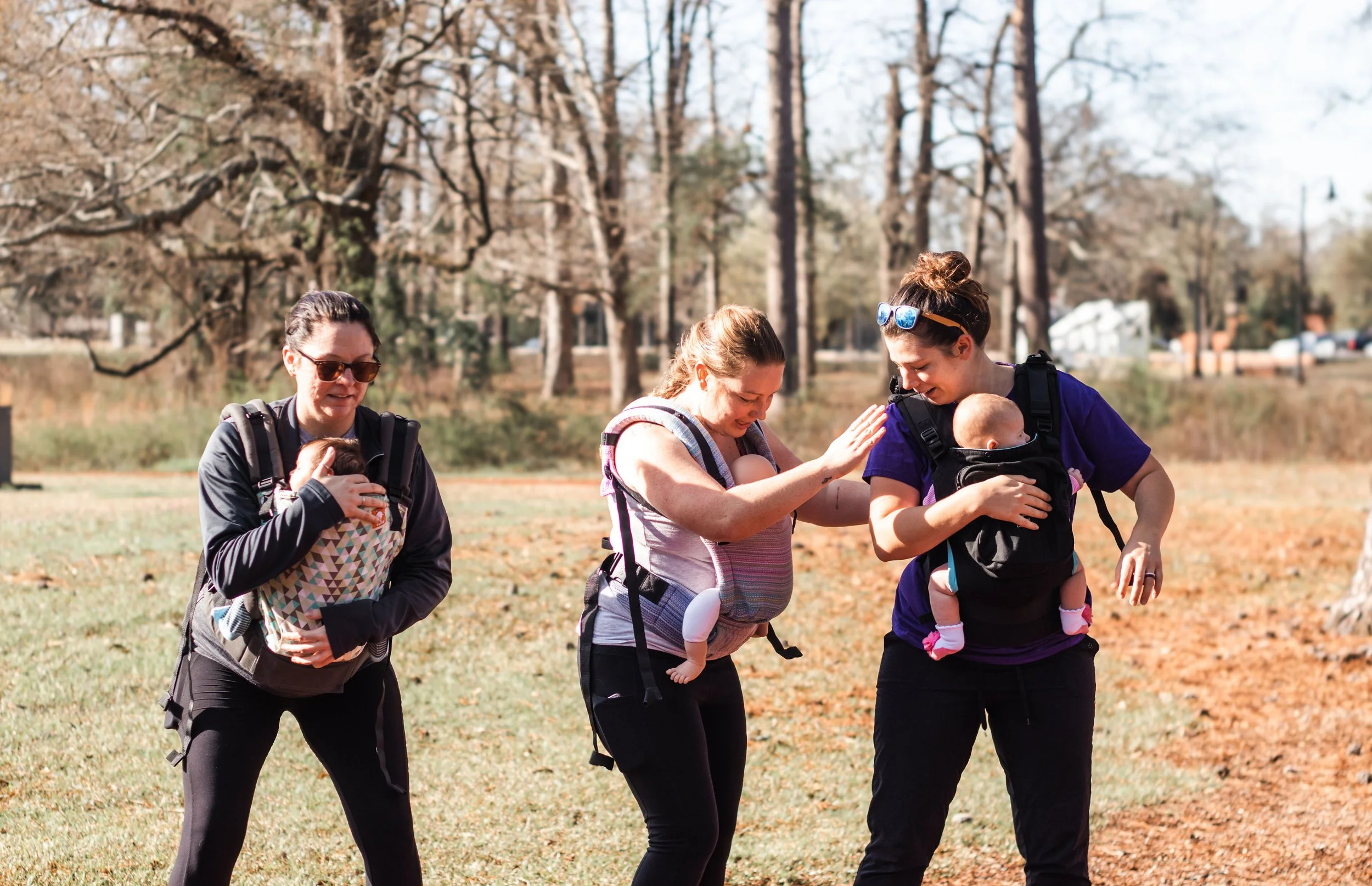 Three women supporting babies in backpacks walk outdoors in a park with leafless trees and grass, enjoying a sunny day.