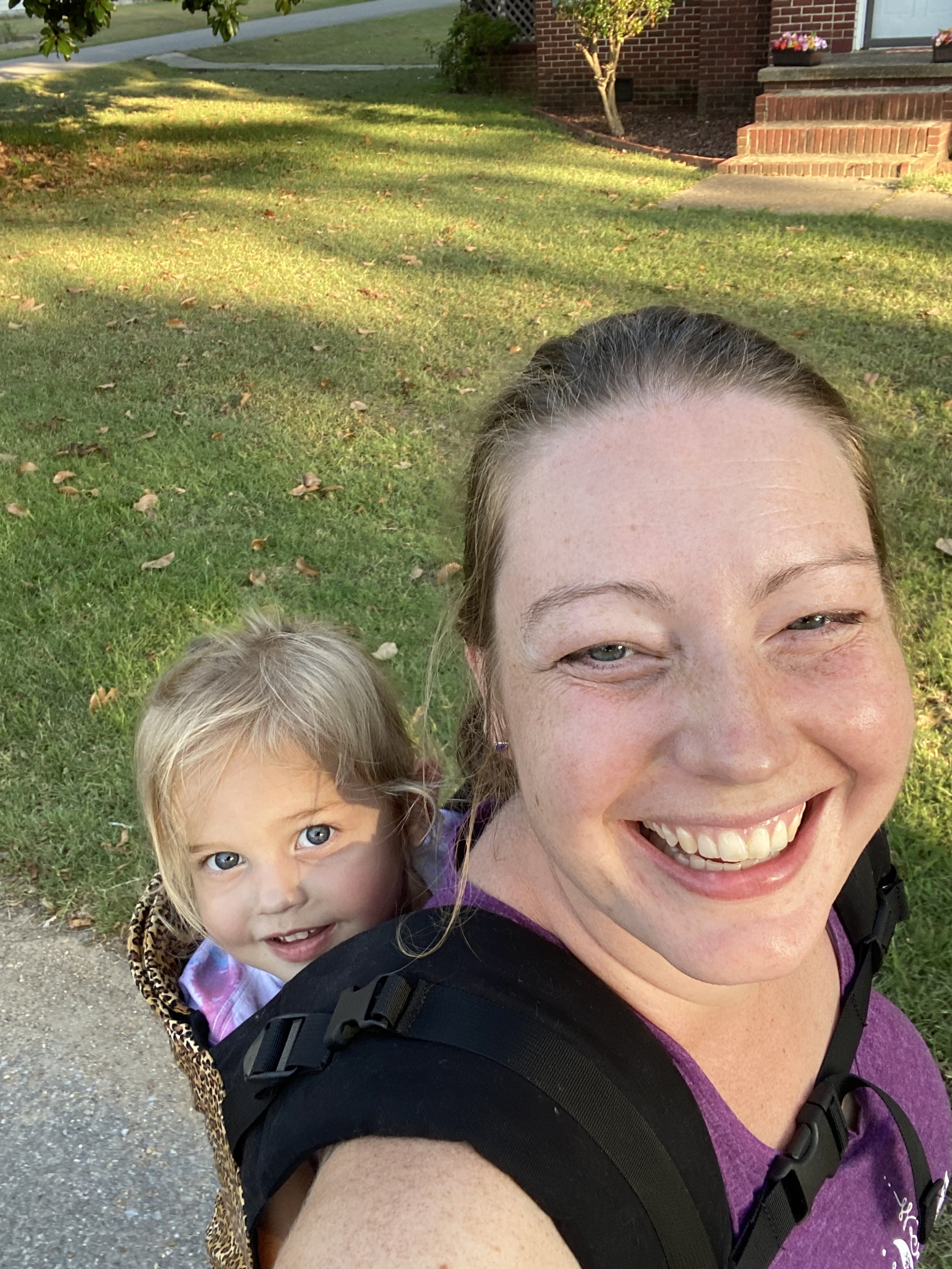 A woman carrying a young girl in a leopard print carrier on her back. The woman is smiling broadly and the girl is looking at the camera with a curious expression. They are outdoors on a sunny day with green grass and a brick house in the background.