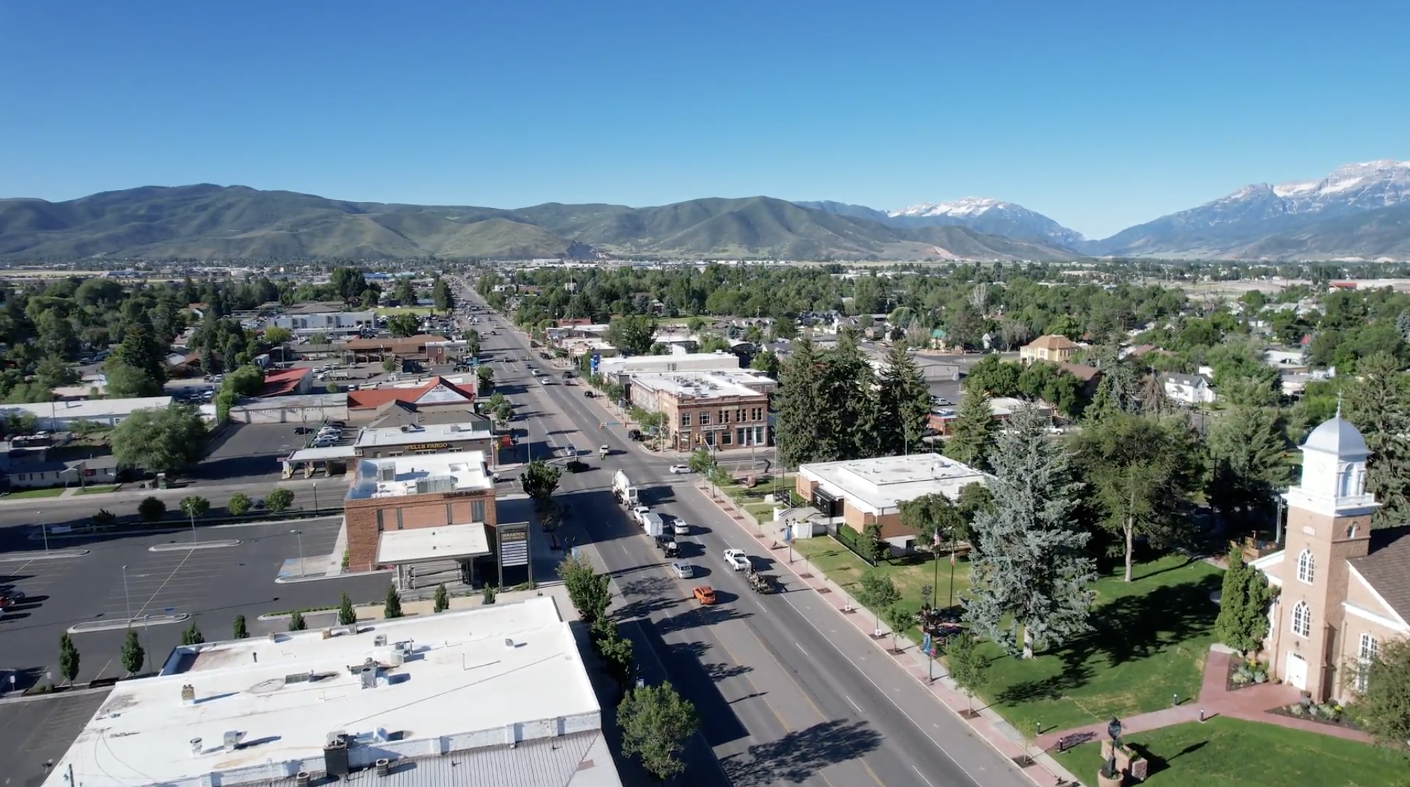 An aerial view of a small town with streets, buildings, and trees, set against a mountainous landscape with snow-capped peaks under a clear blue sky.