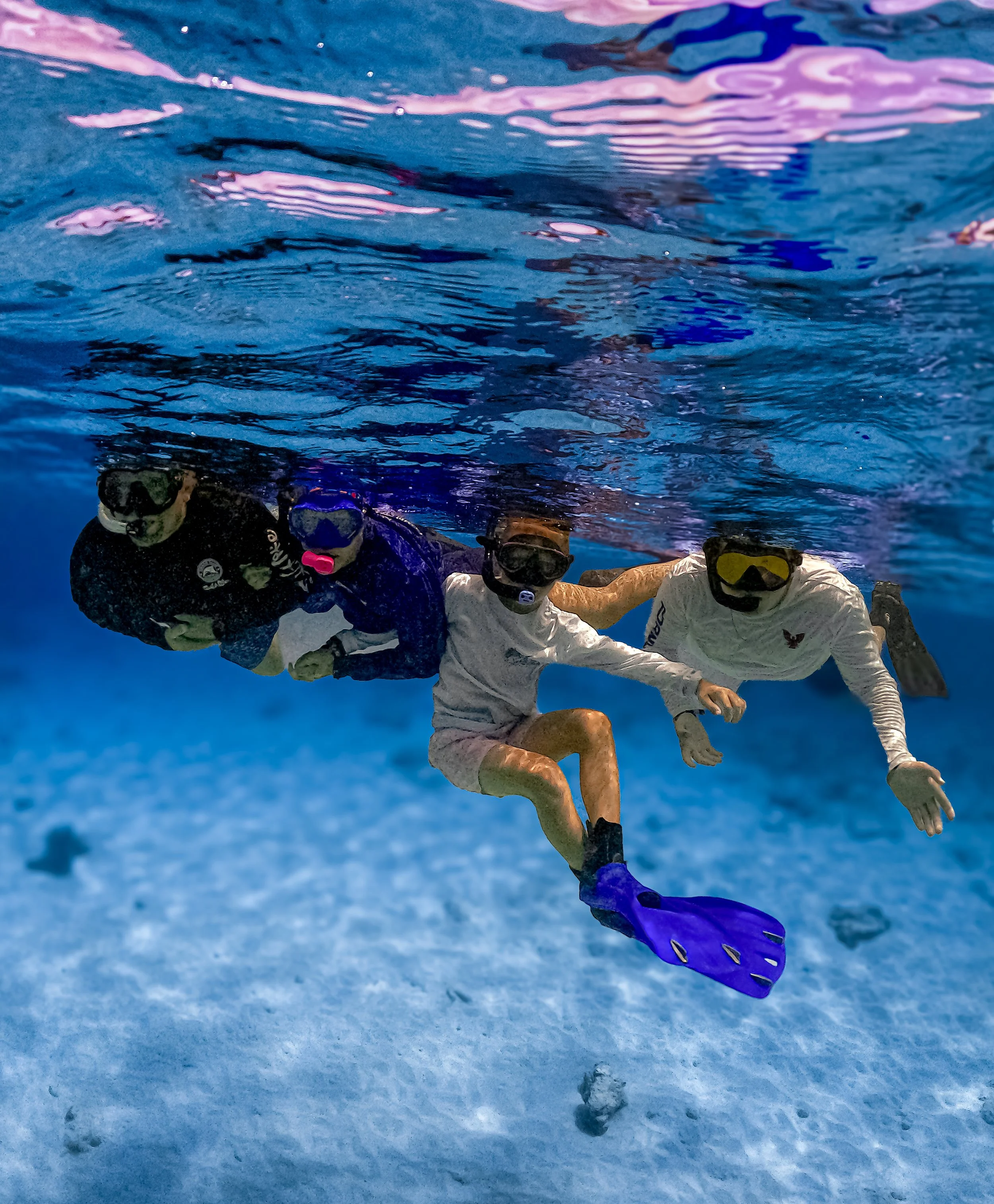 A group of people underwater wearing snorkeling gear, holding hands, and swimming in a line, with one person holding a purple fins, in a body of water with a blue tint.