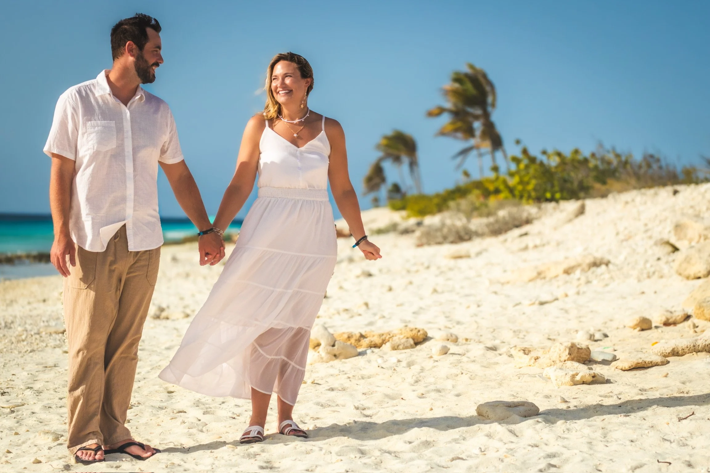 A smiling man and woman hold hands while walking on a sandy beach with palm trees and turquoise ocean in the background.