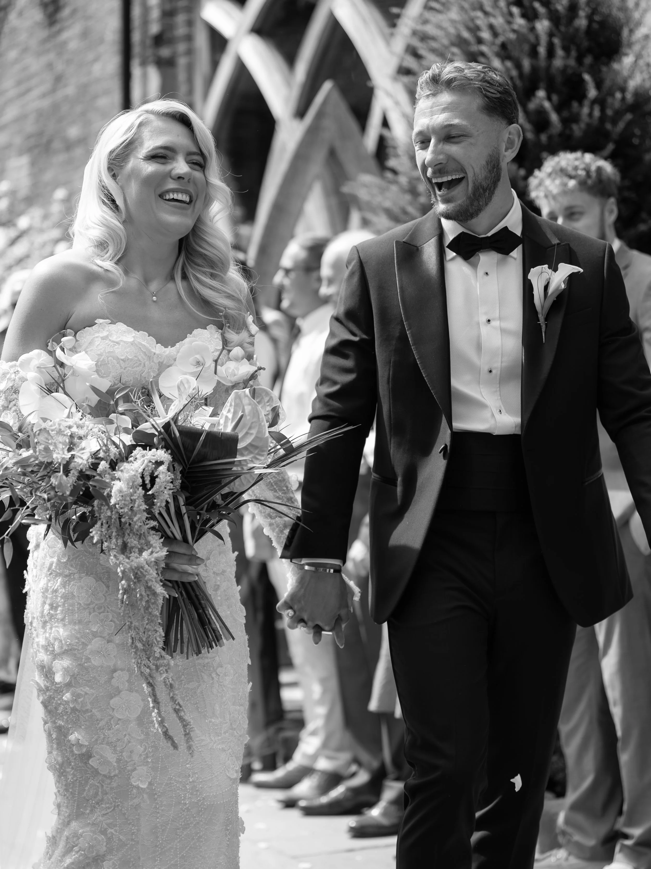 Black and white photo of a bride and groom holding hands and smiling during their wedding ceremony, with guests in the background.
