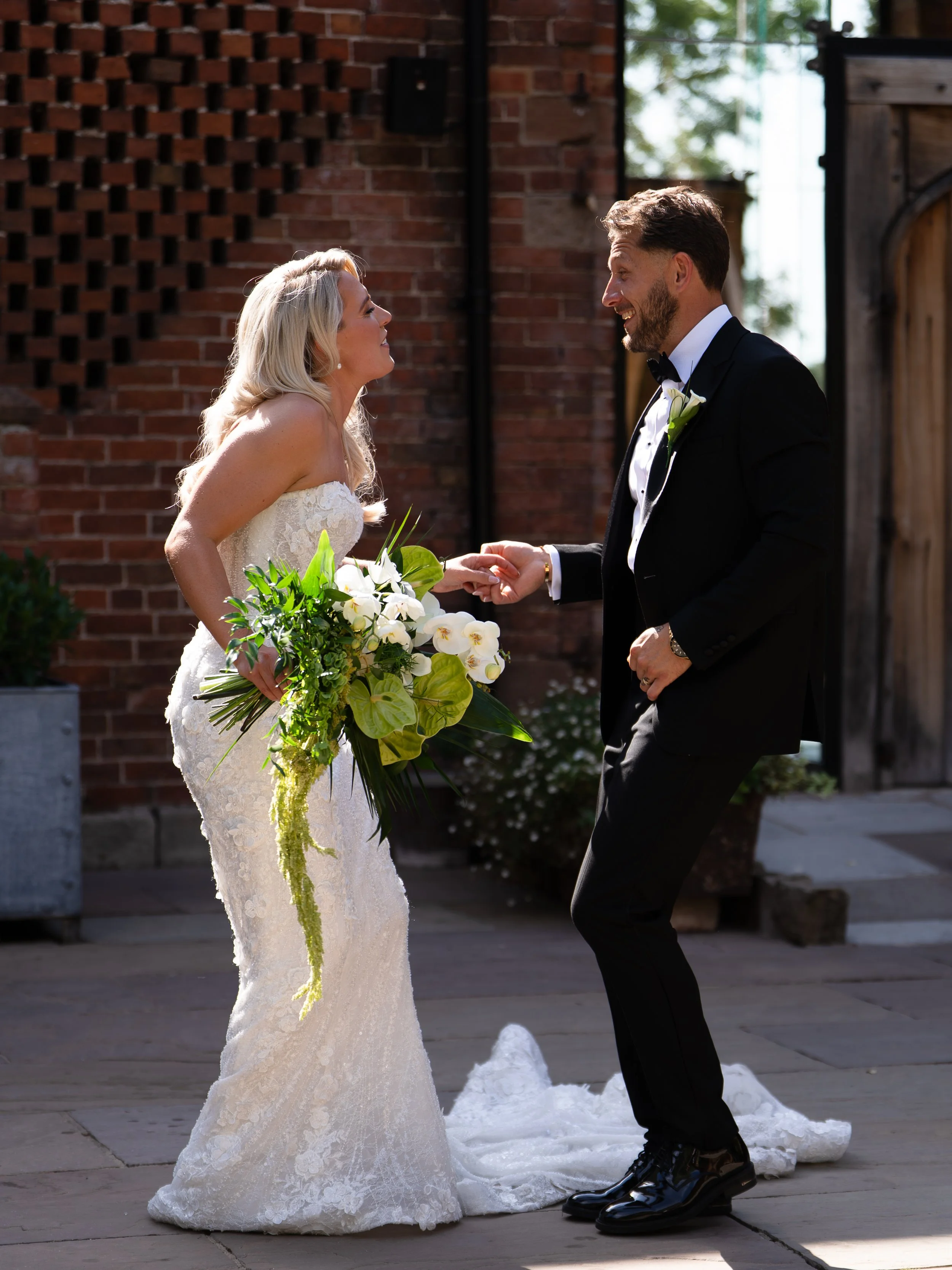 A bride and groom hold hands during their wedding ceremony outdoors, with a brick wall and wooden gate in the background. The bride wears a lace wedding dress and holds a bouquet of white flowers and green foliage. The groom wears a black tuxedo with a white shirt and black bow tie.