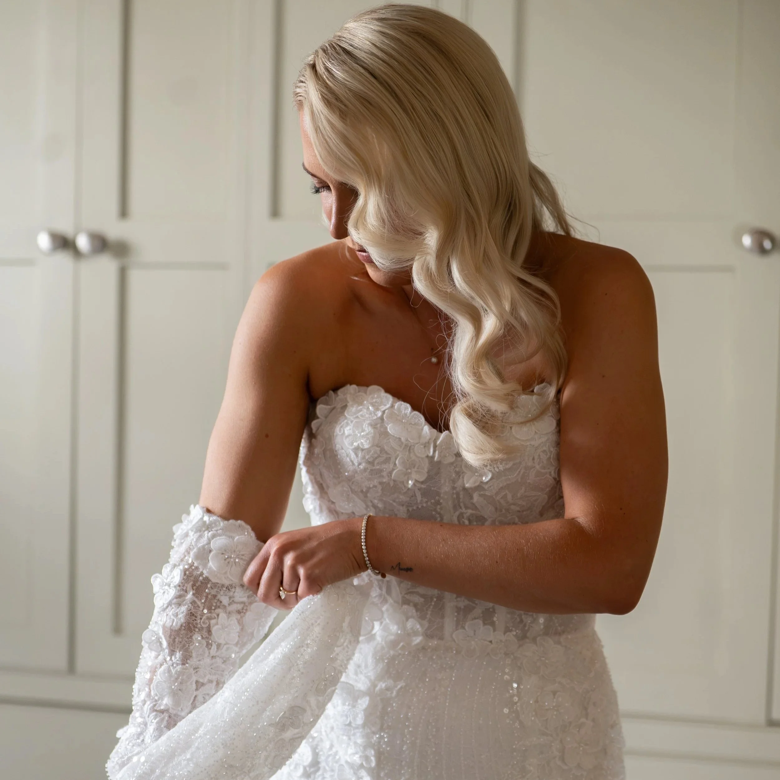 A woman with long blonde hair in loose curls wearing a lace strapless wedding dress with long sleeves - Keira by Blanche Bridal at Victoria Lou Bridal. 