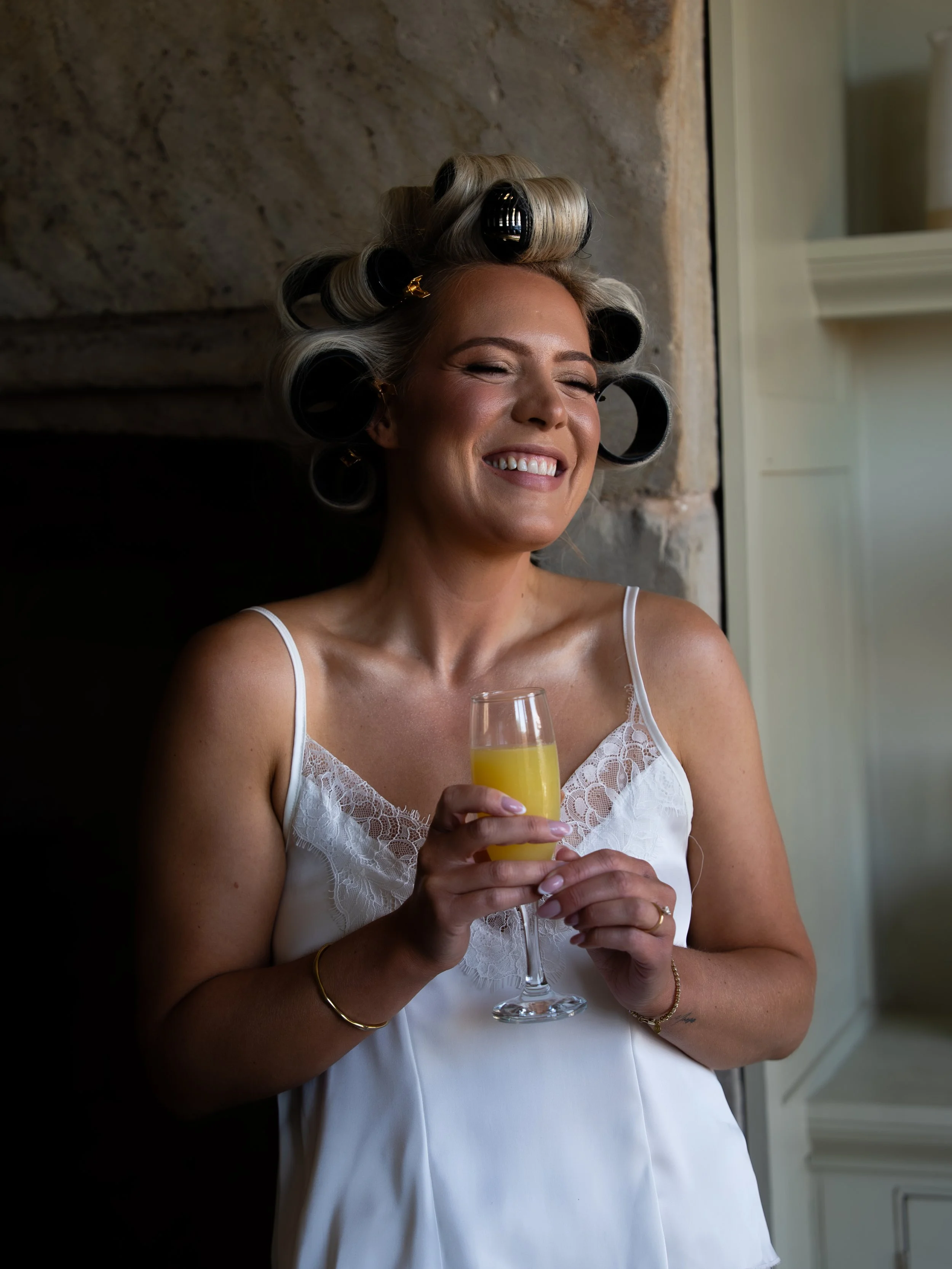 Woman with hair rollers in her hair, wearing a white lace top, smiling and holding a glass of mimosa near a stone wall and a window.