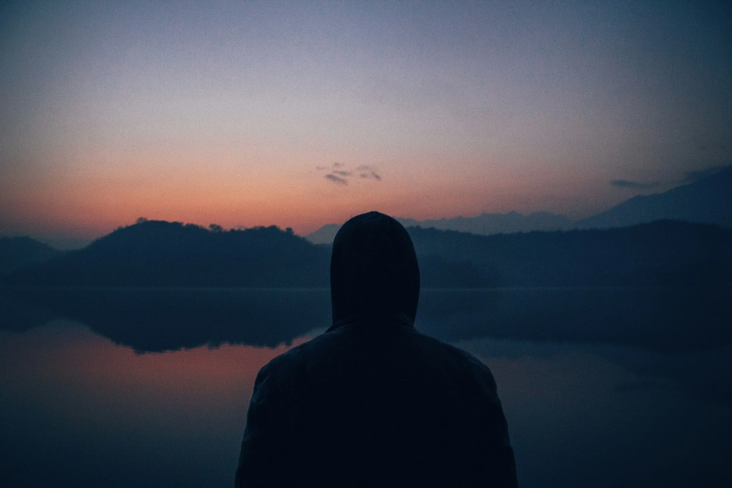 Person sitting by a lake during sunset, surrounded by mountains.