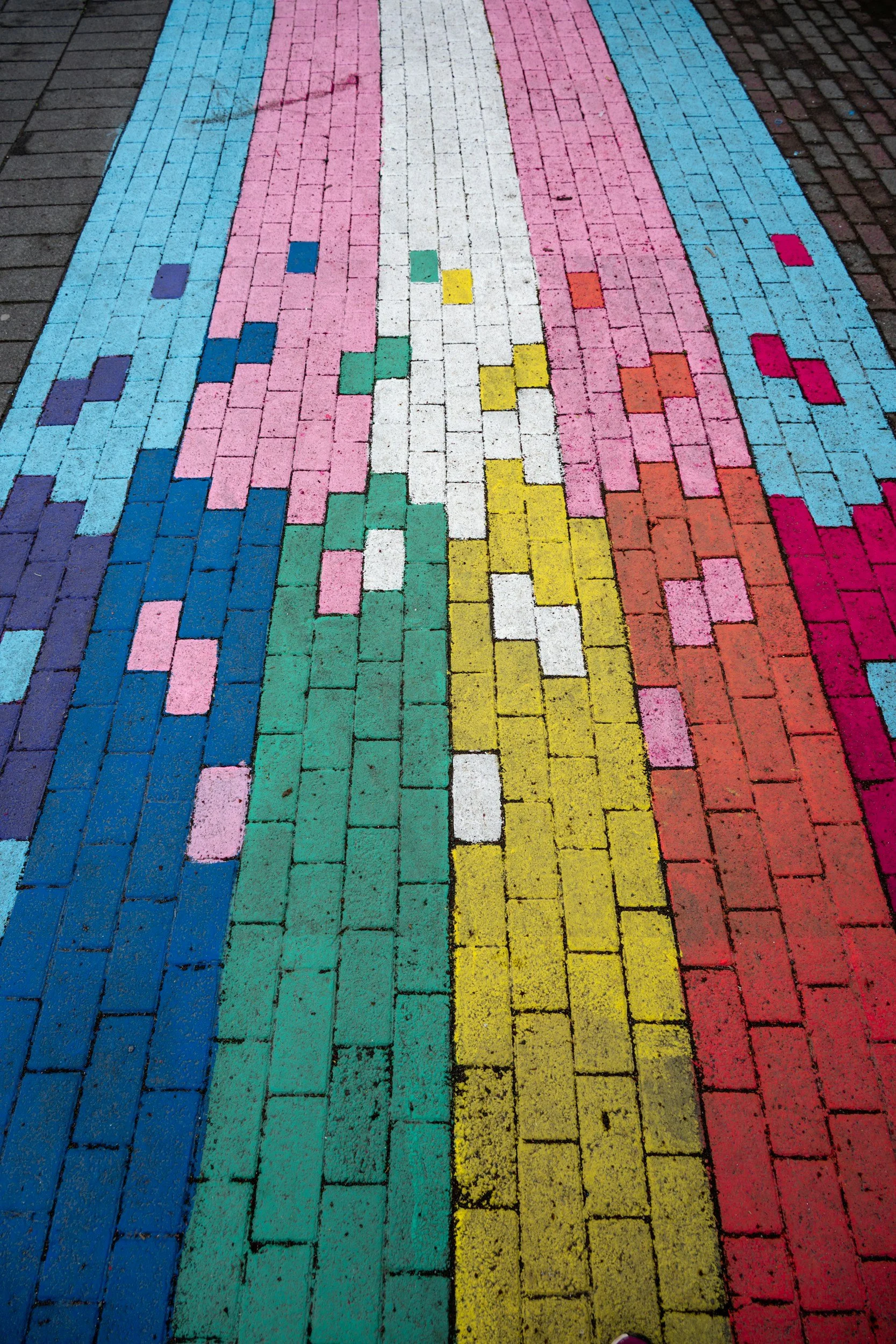 Colorful rainbow pattern painted on brick pavement with vertical stripes in blue, pink, white, yellow, orange, red, and purple.