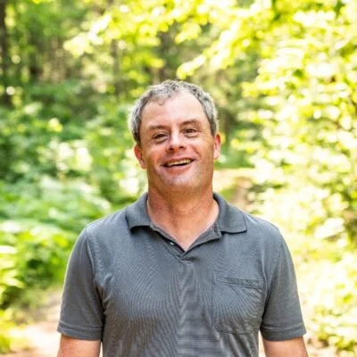 A smiling man standing outdoors in front of green trees, wearing a gray polo shirt.