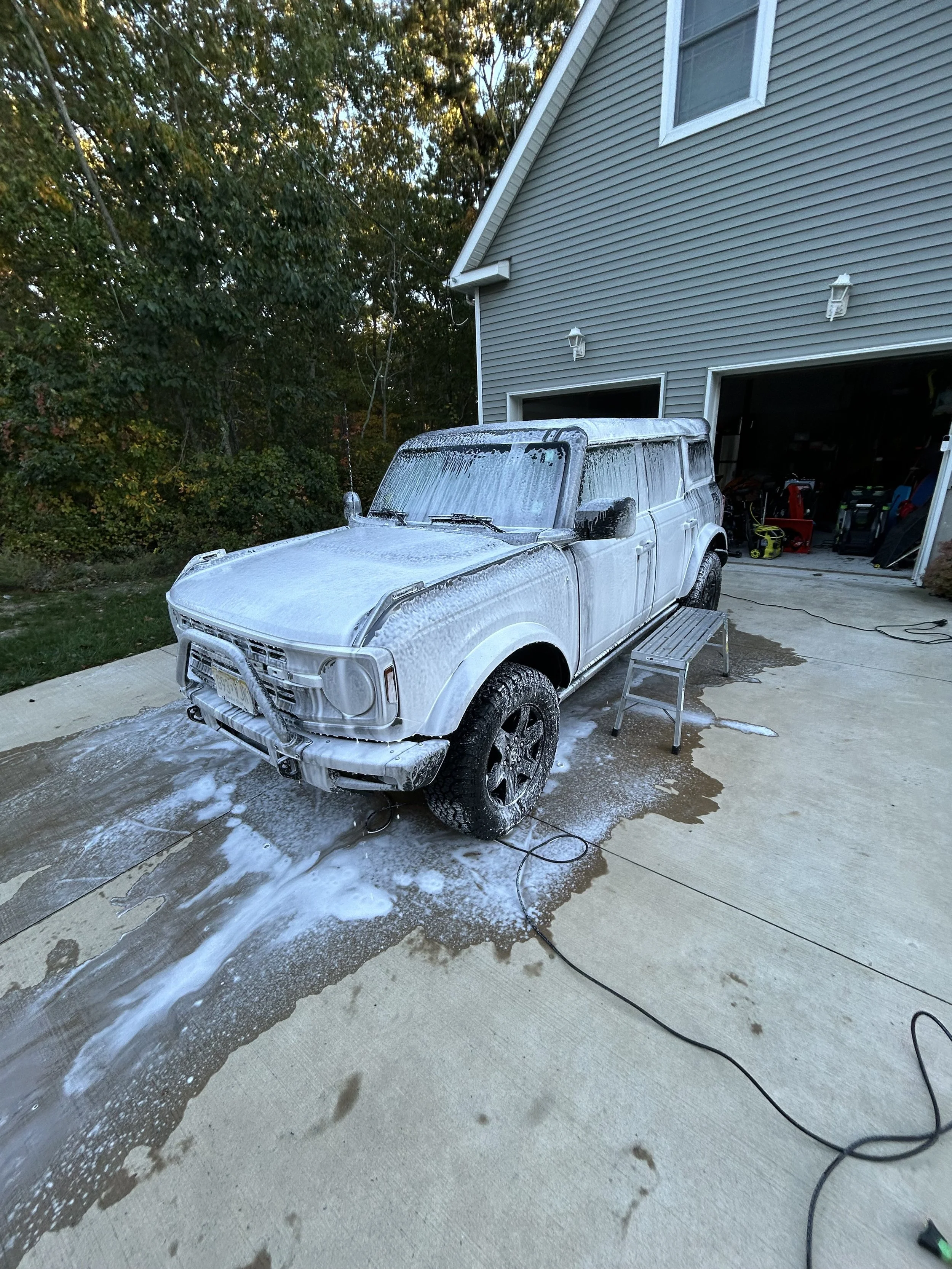 A white SUV being washed with soap on a concrete driveway in front of a gray house with an open garage. The SUV is covered in soap with foam on the windshield and hood. A small step stool is beside the vehicle, and there are cleaning supplies and tools in the garage.