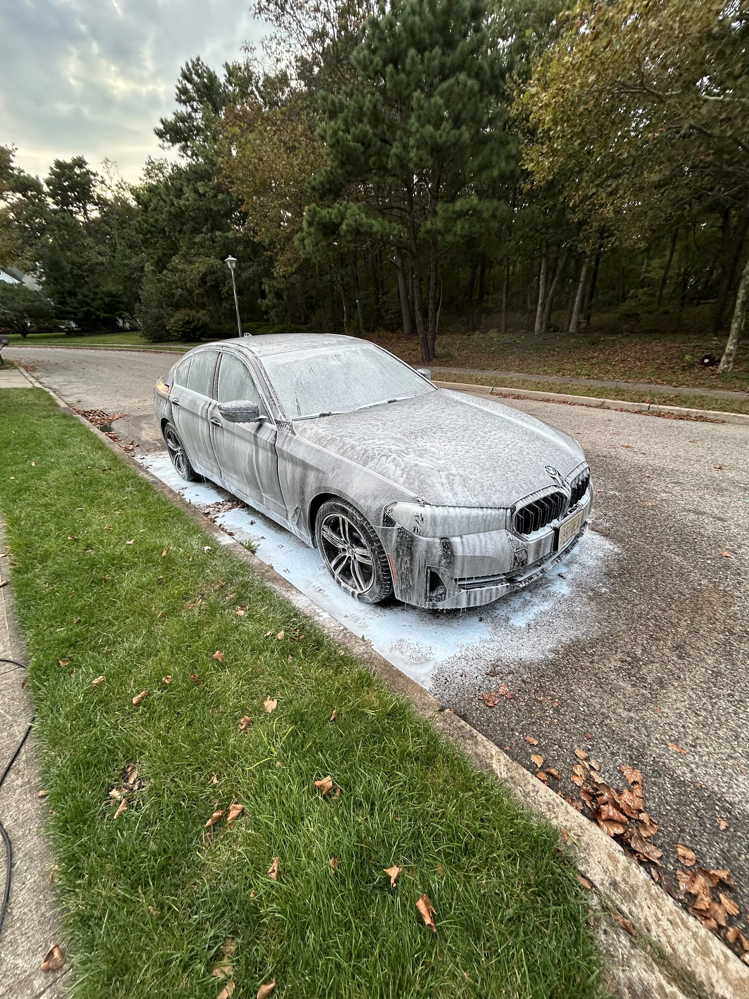 A black BMW sedan covered in soap suds parked on a residential street with green grass and trees in the background.