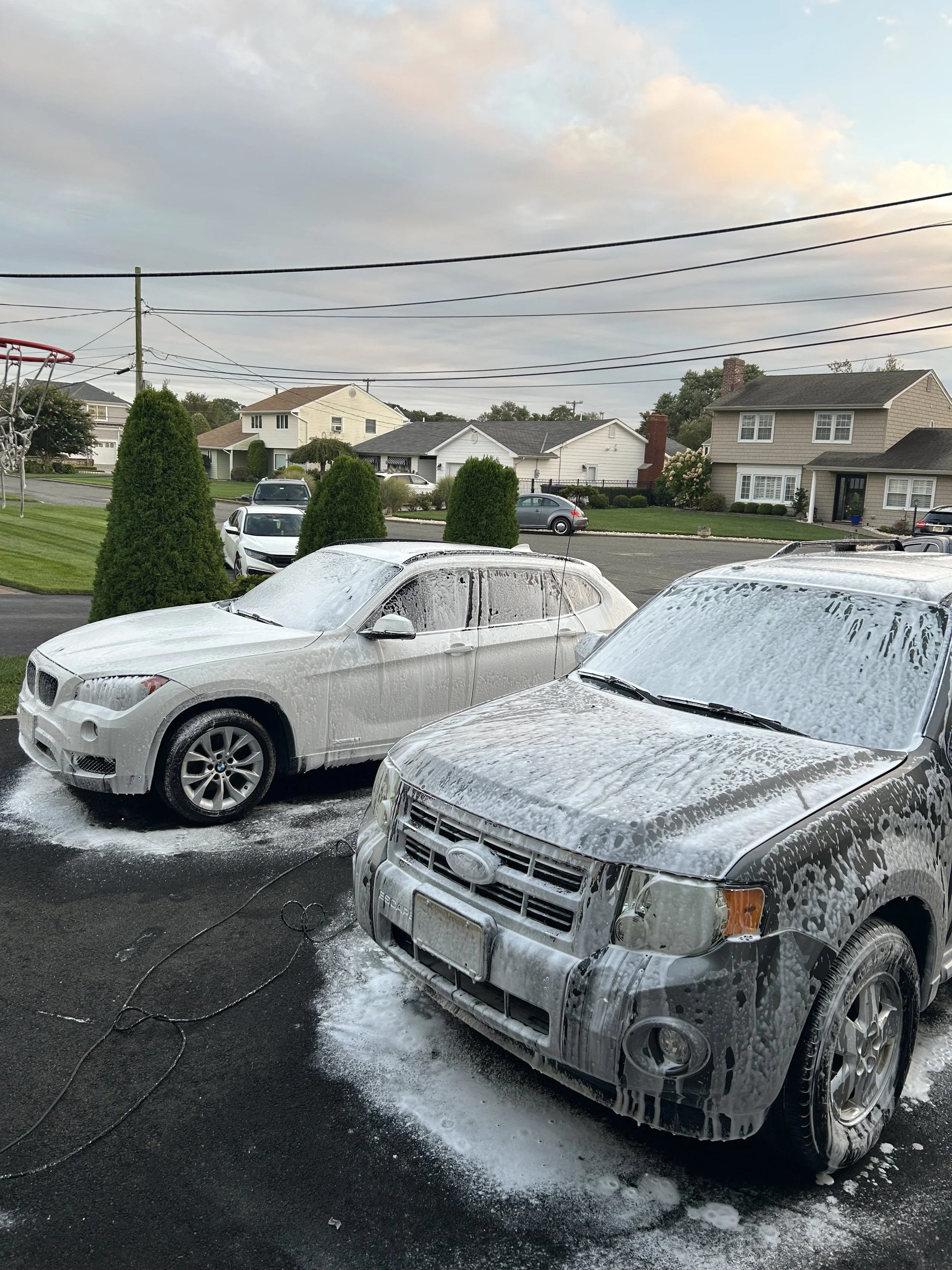 Two silver cars are undergoing a car wash with soap and foam on the hood, windshield, and roof, with water and soap suds on the ground. The background shows a residential neighborhood with houses, green lawns, and trees, under a partly cloudy sky.