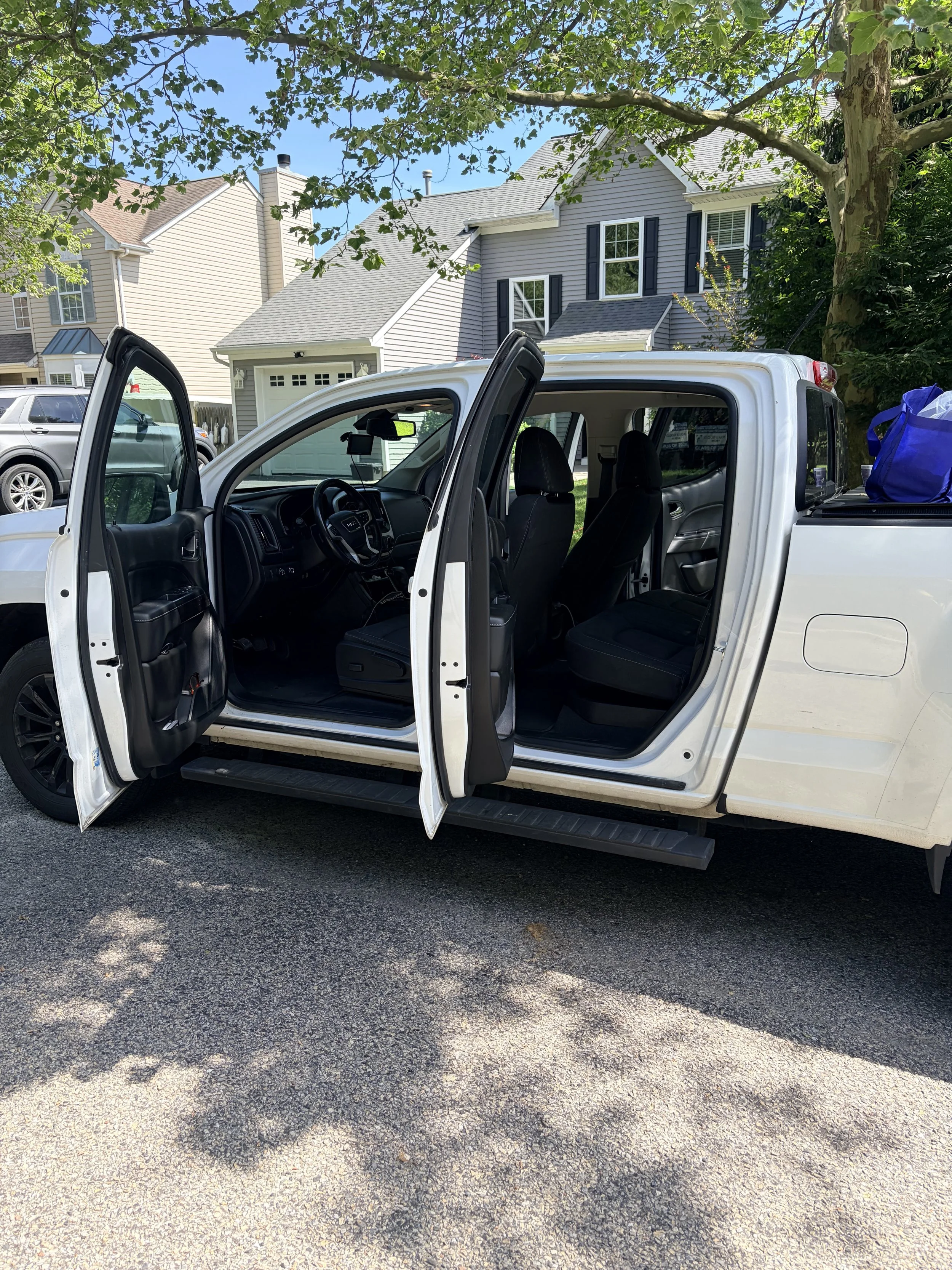 A white pickup truck with its front and rear passenger side doors open, parked on a paved street in front of a residential neighborhood with houses, trees, and other parked cars. The truck's interior is visible, and a blue bag is in the truck bed.