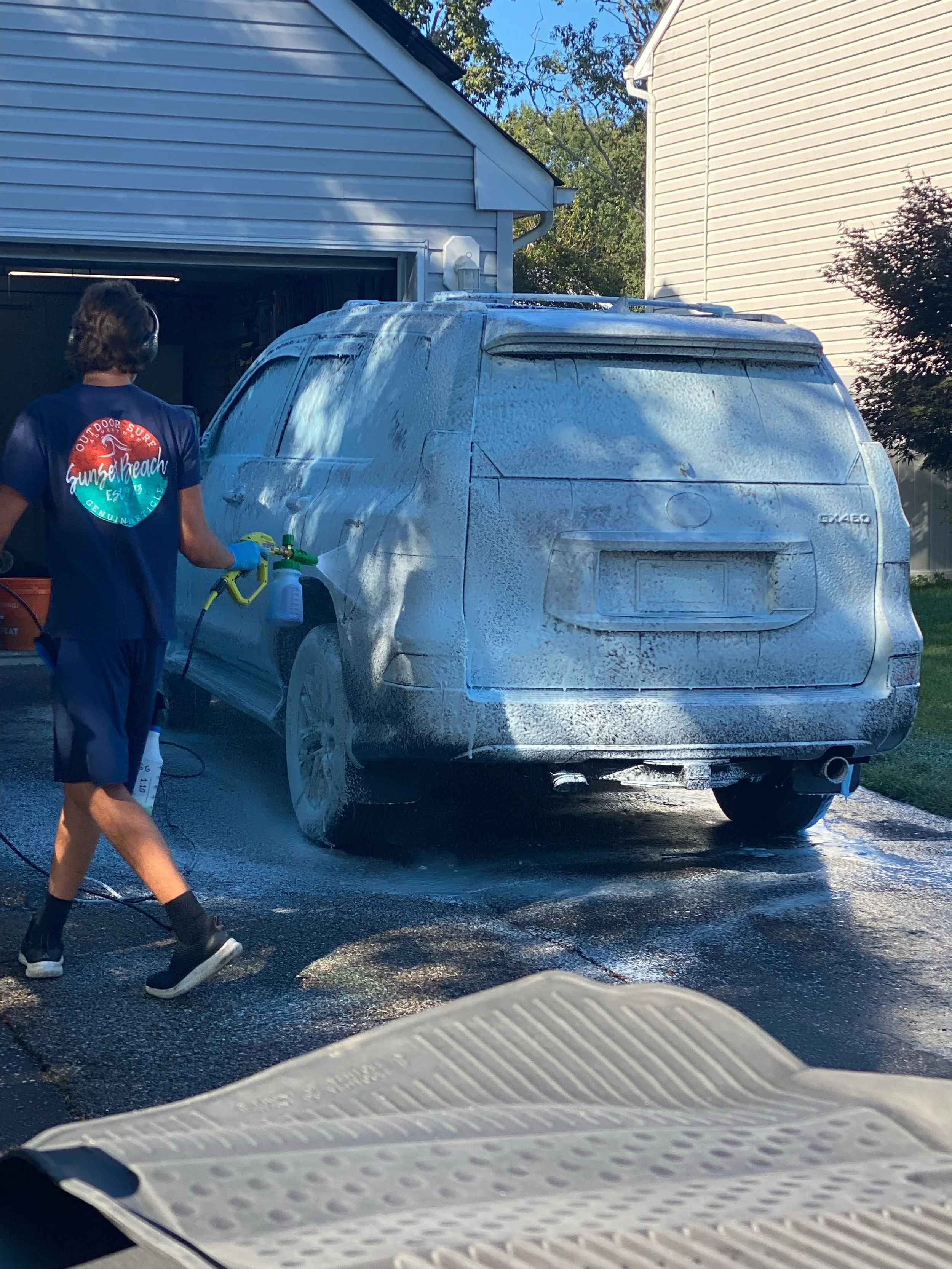 A person wearing a blue t-shirt, shorts, and headphones is pressure washing a silver SUV covered in blue foam, in a driveway next to a house with white siding.