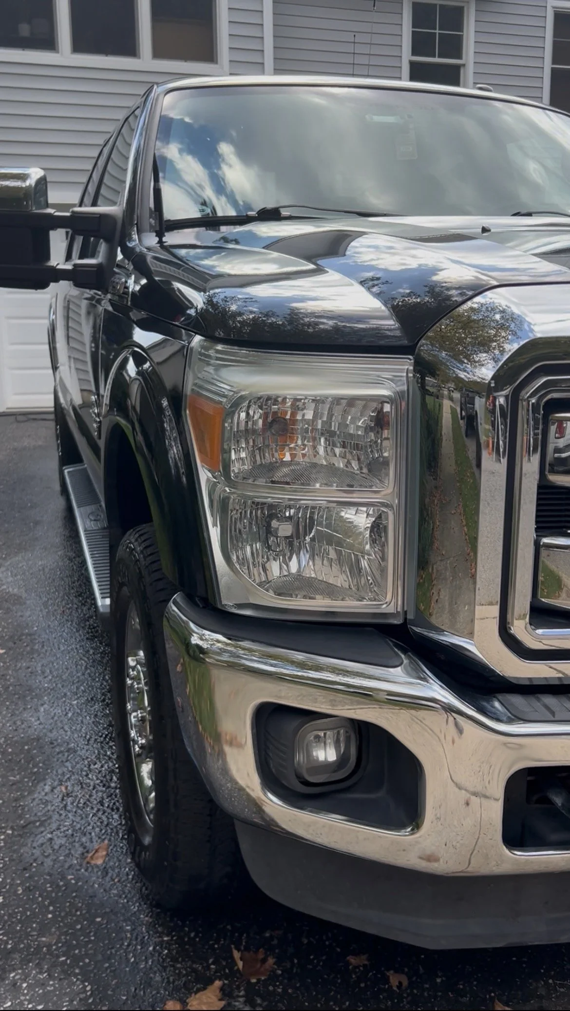 Close-up of the front part of a black and chrome pickup truck parked on an asphalt driveway with fallen leaves, house siding, and windows reflected on the vehicle's surface.