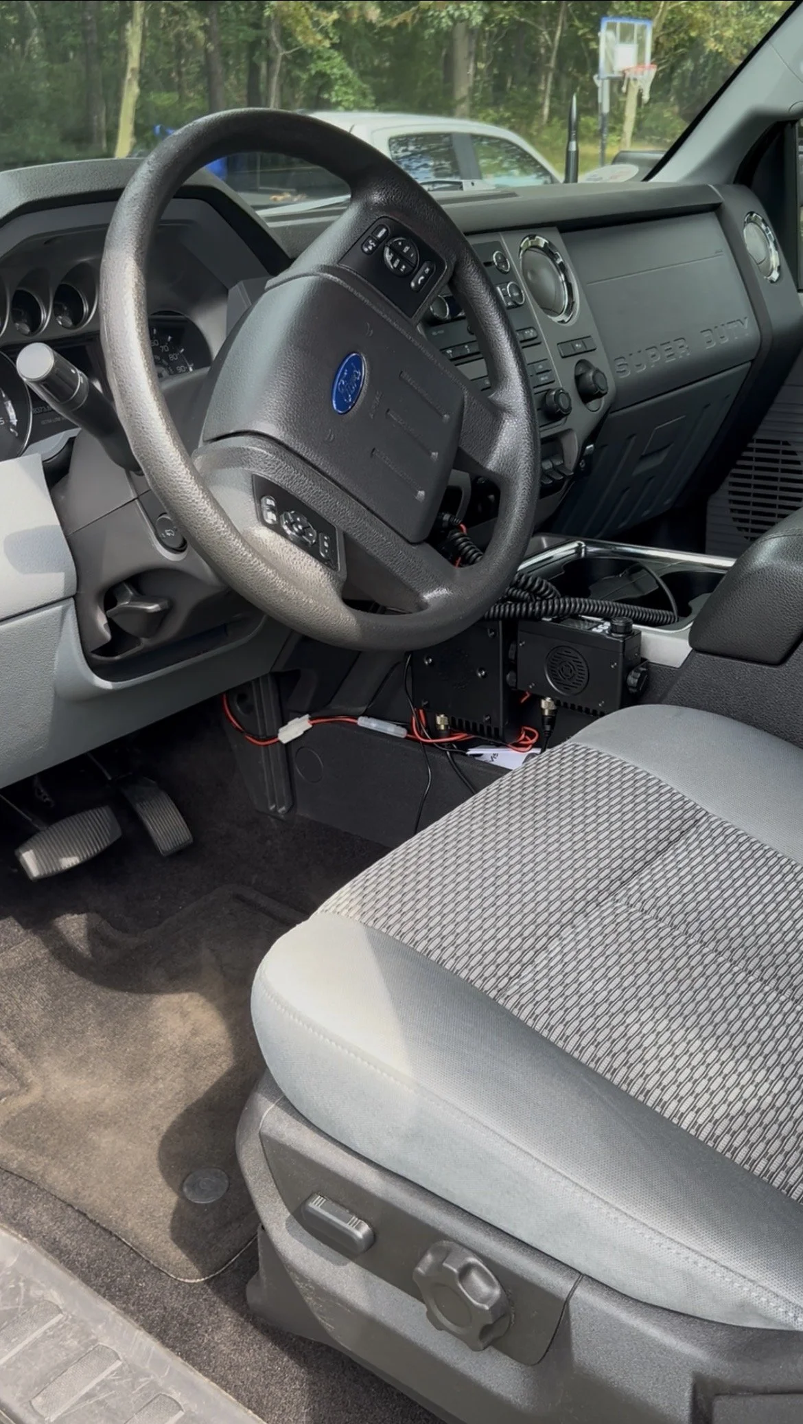 Interior of a Ford Super Duty truck with a steering wheel, dashboard, and driver's seat, featuring a mounted electronic device with wires underneath and a view of a wooded area outside.