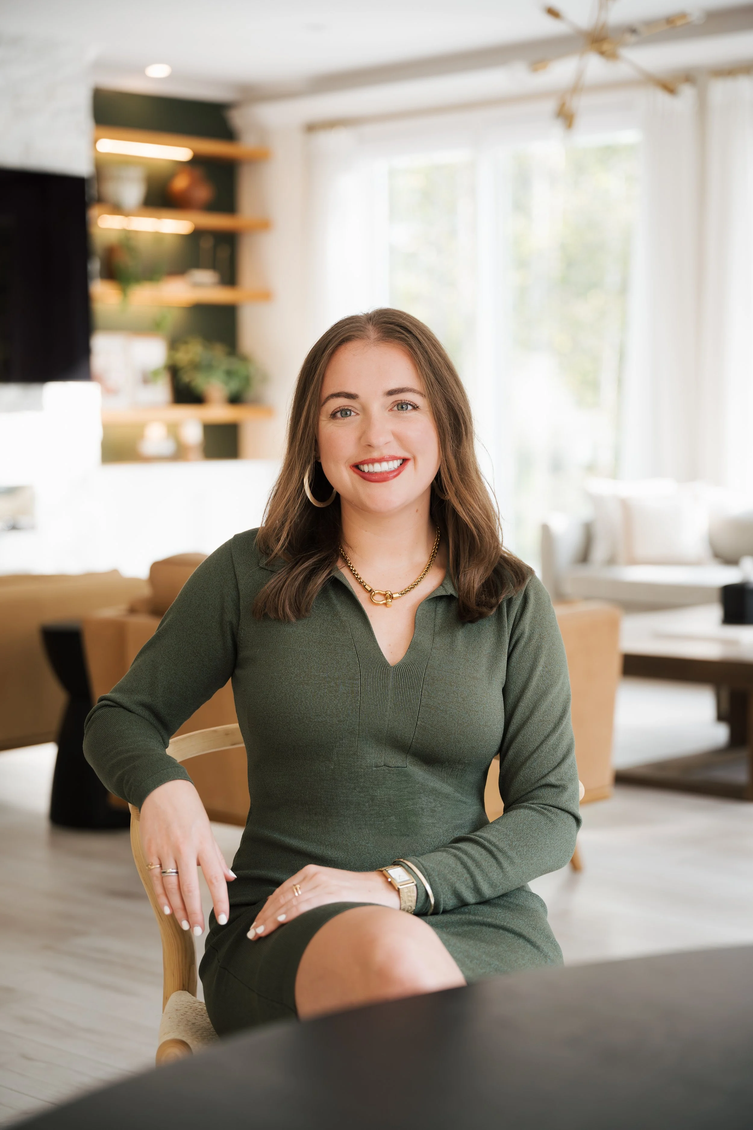A woman sitting on a chair in a cozy, well-lit living room with large windows, wearing a green top, smiling, with jewelry and a watch, with a blurred foreground.