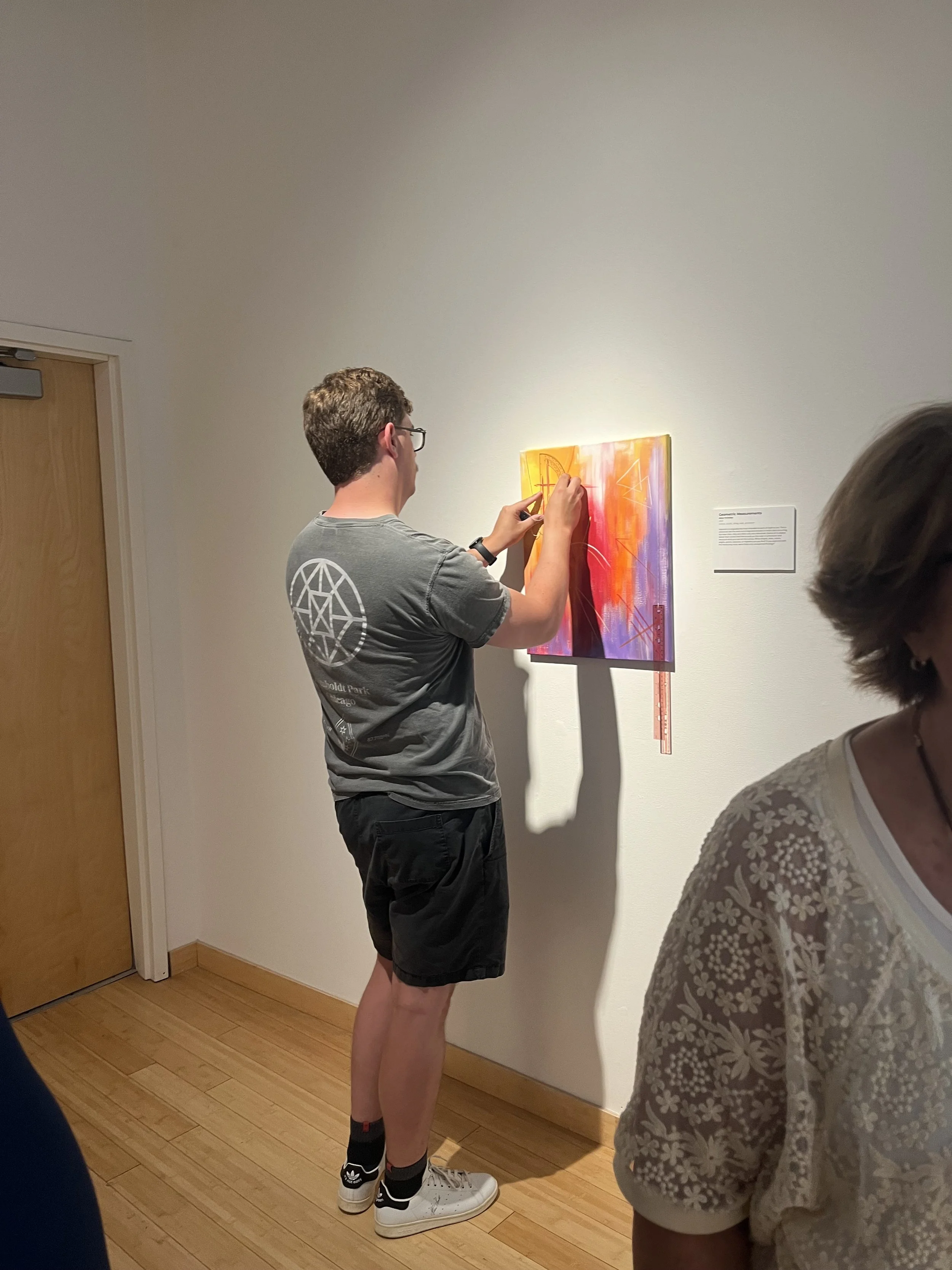 A young man wearing glasses, a gray graphic t-shirt, black shorts, and white sneakers is drawing on a colorful abstract painting with a pencil in an art gallery. There are other people with similar minimalistic furnishings in the gallery.
