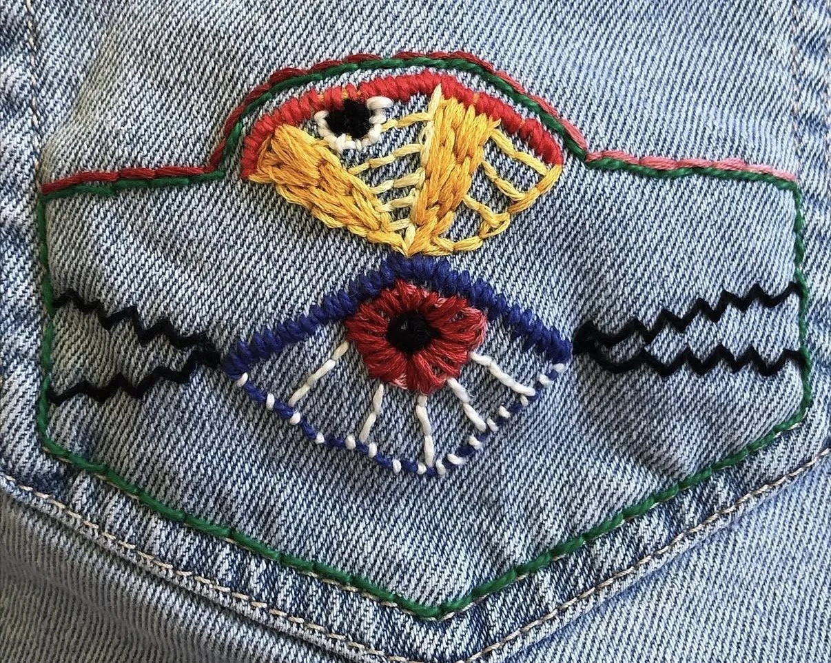 An embroidered patch of an eye with a yellow butterfly on the upper eyelid, a red heart in the center, and black and white details, on denim fabric.
