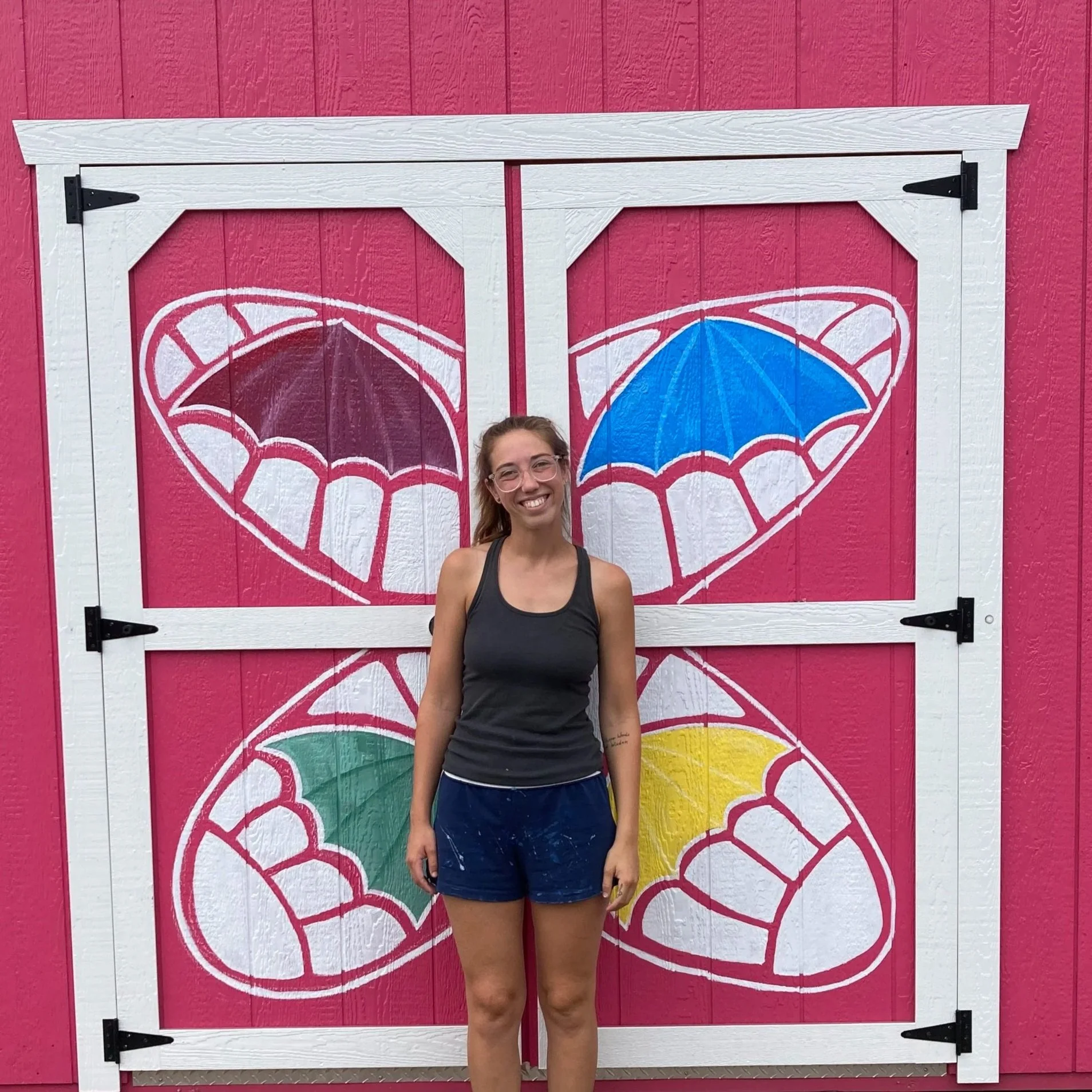 Young woman with glasses standing in front of a pink wall with large butterfly murals and a white frame.