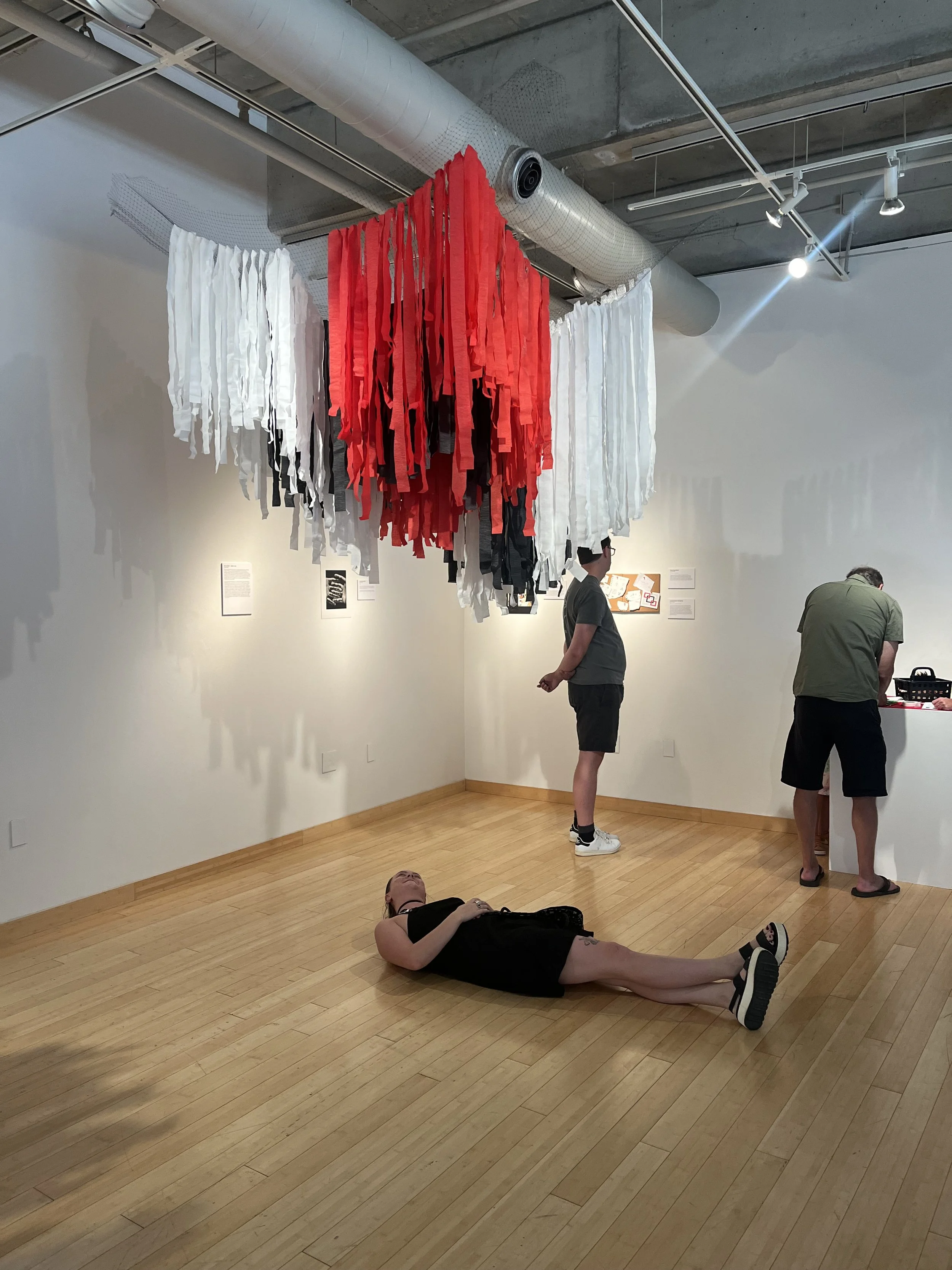 An art gallery or museum room with abstract hanging fabric art consisting of red, black, and white strips suspended from the ceiling. Several visitors are seen: a woman lying on the wooden floor, a woman standing and looking at her phone, and two oth