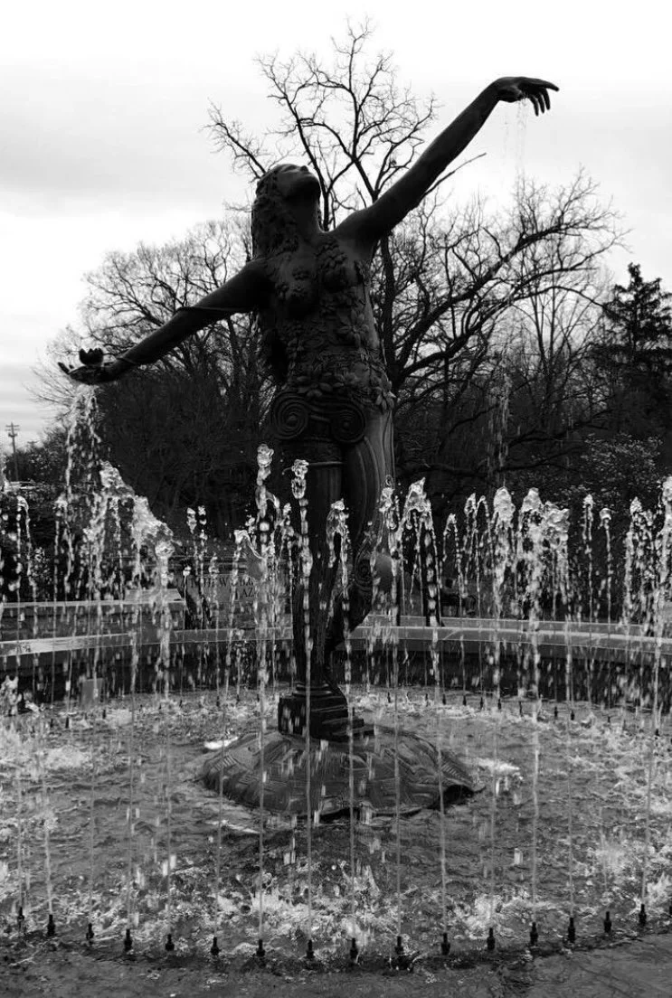 Black and white photo of a statue of a woman with arms outstretched, standing on a fountain with water jets around her, and leafless trees in the background.