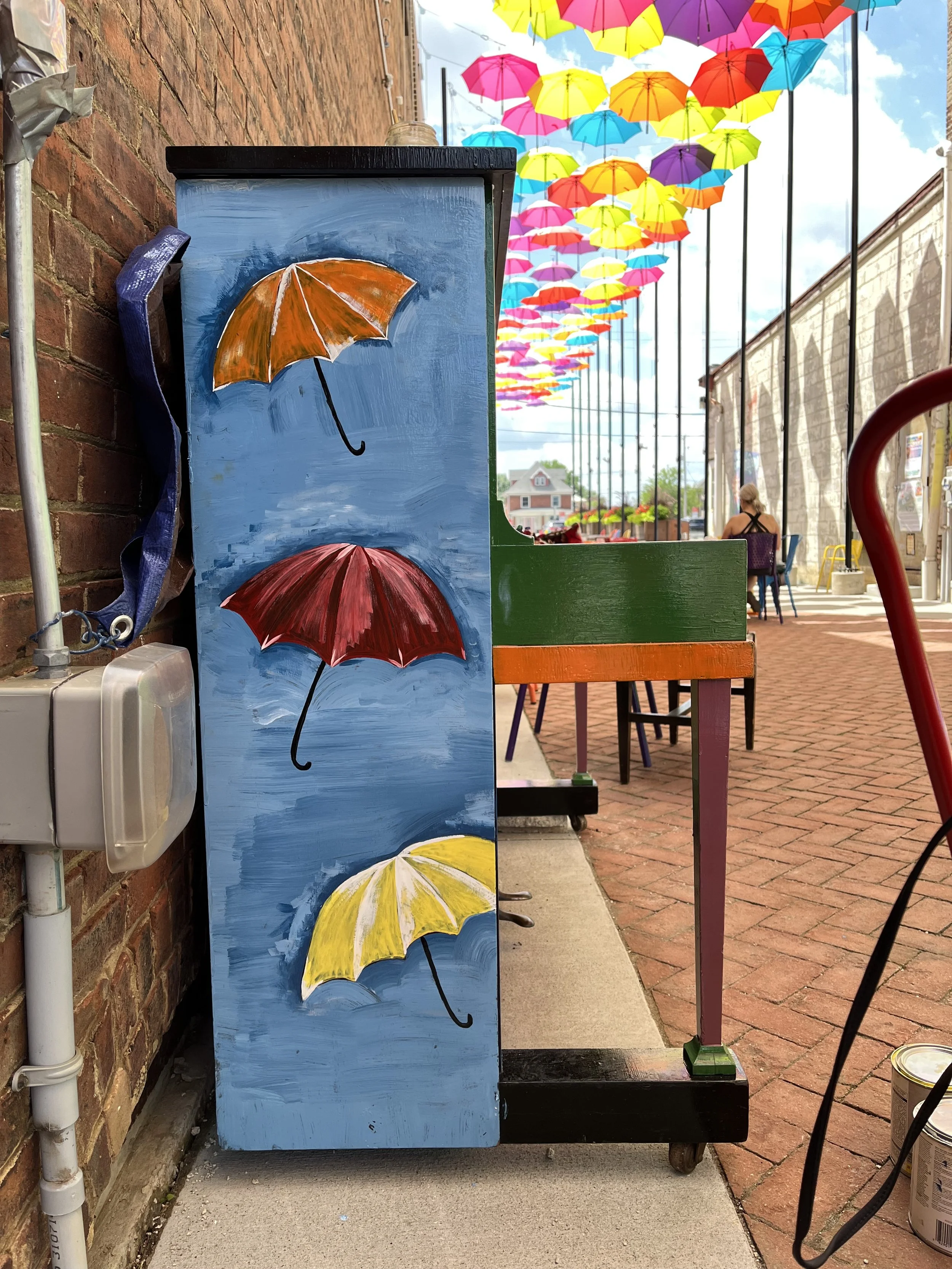 A colorful outdoor art display depicts umbrellas painted on the side of a blue box, with additional umbrellas hanging overhead in bright colors like pink, yellow, and purple, against a partly cloudy sky and brick sidewalk.