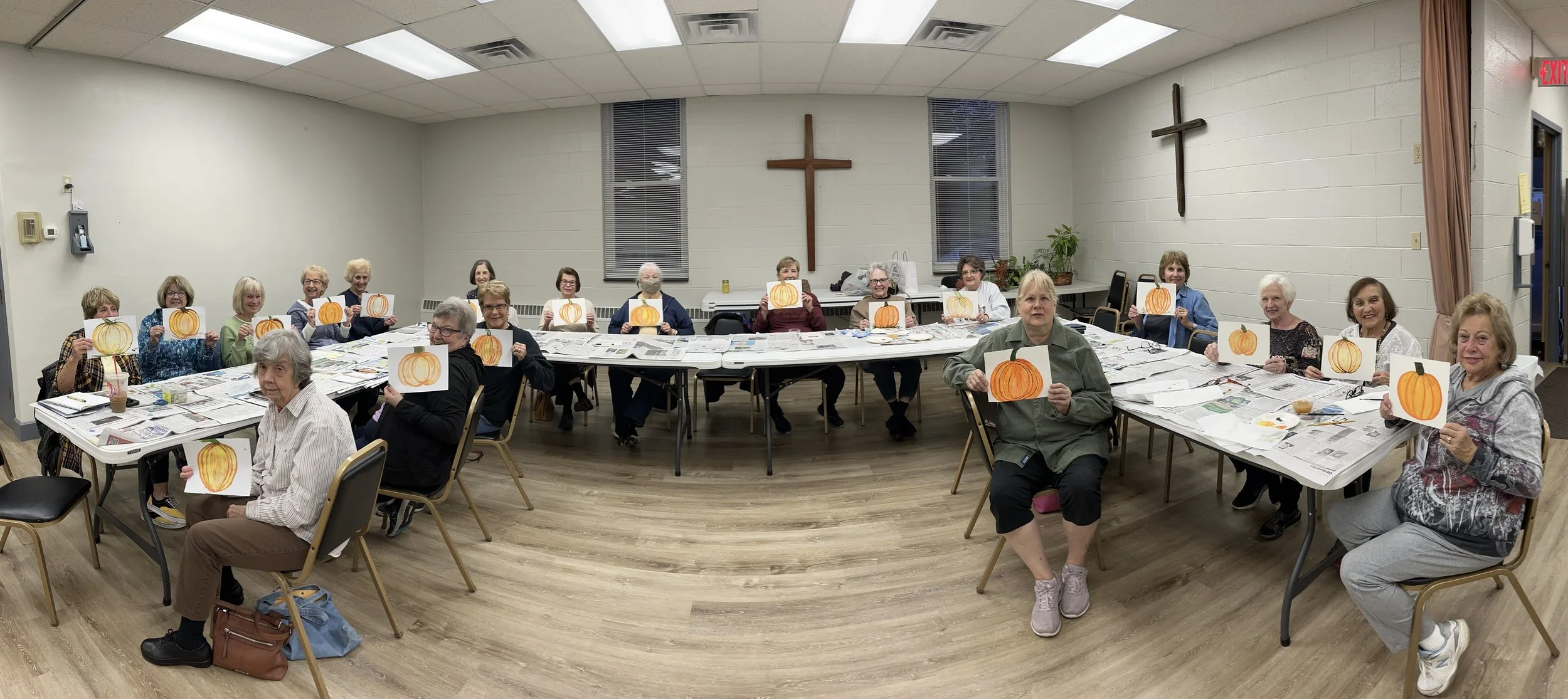 Group of elderly women sitting around a large table in a community room, each holding up a paper with a painted pumpkin, with crosses on the wall behind them.