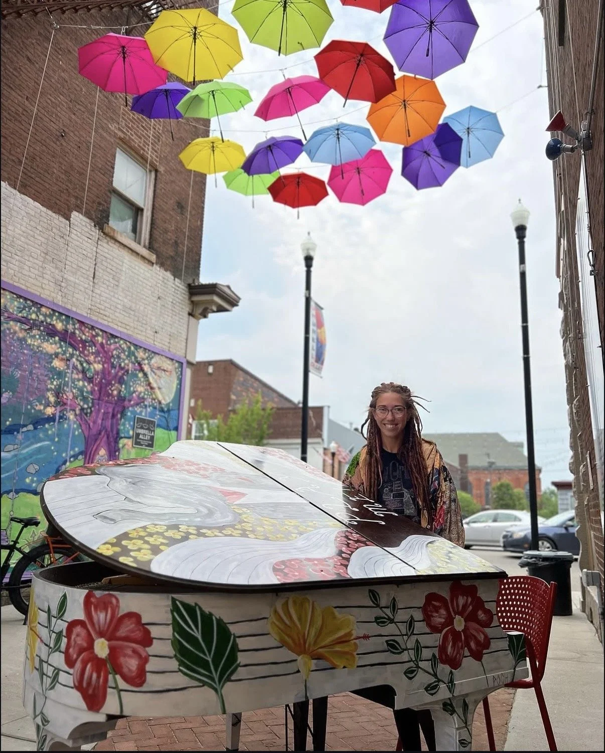 A woman with braids and glasses standing outdoors under colorful umbrellas hanging above. She is smiling and standing behind a decorated painted piano with floral designs, in an urban alley with brick buildings and streetlights.