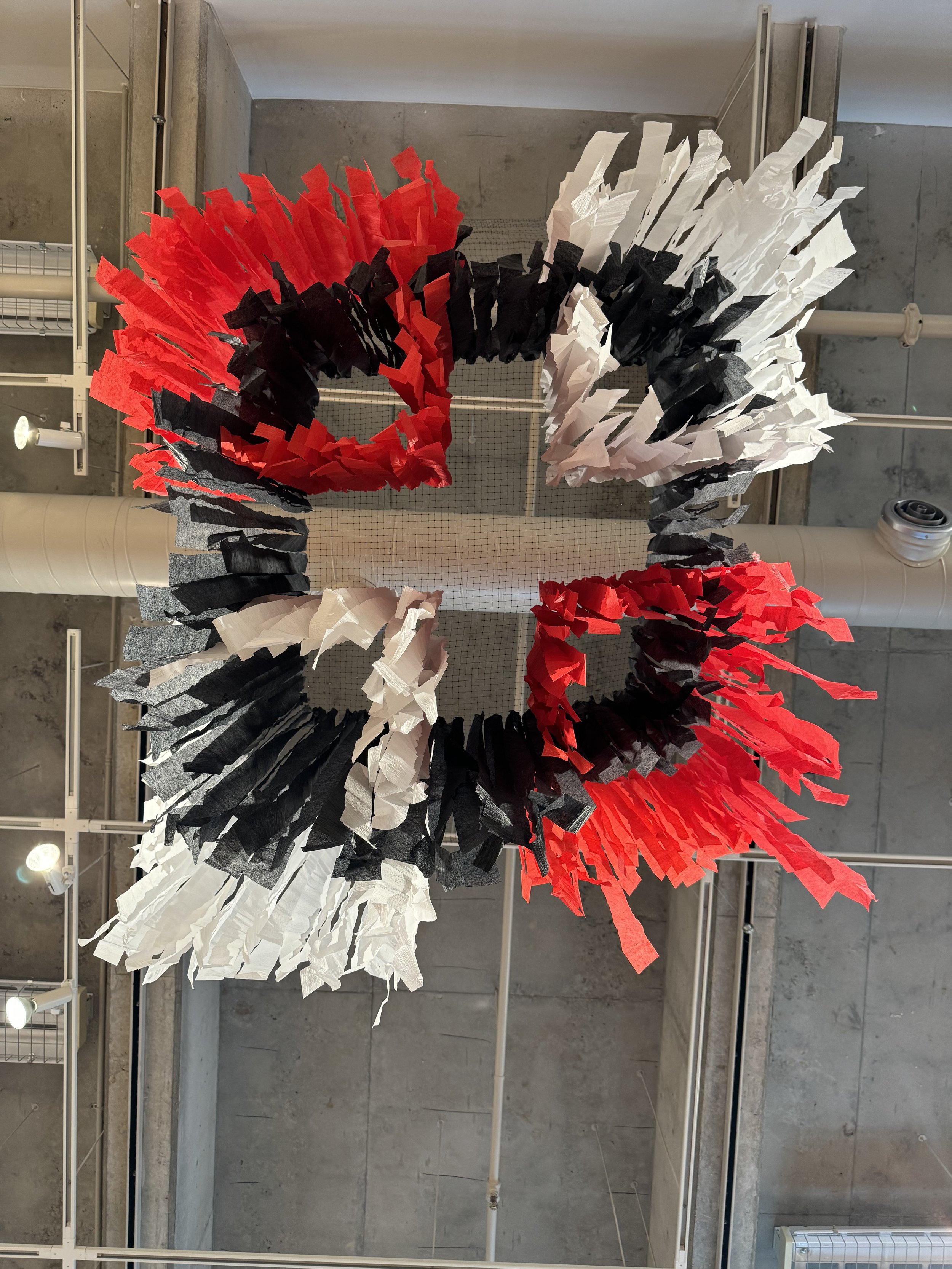 Art installation of a circular wreath made from torn pieces of red, black, white, and gray paper, displayed on a gallery wall with concrete and metal accents.