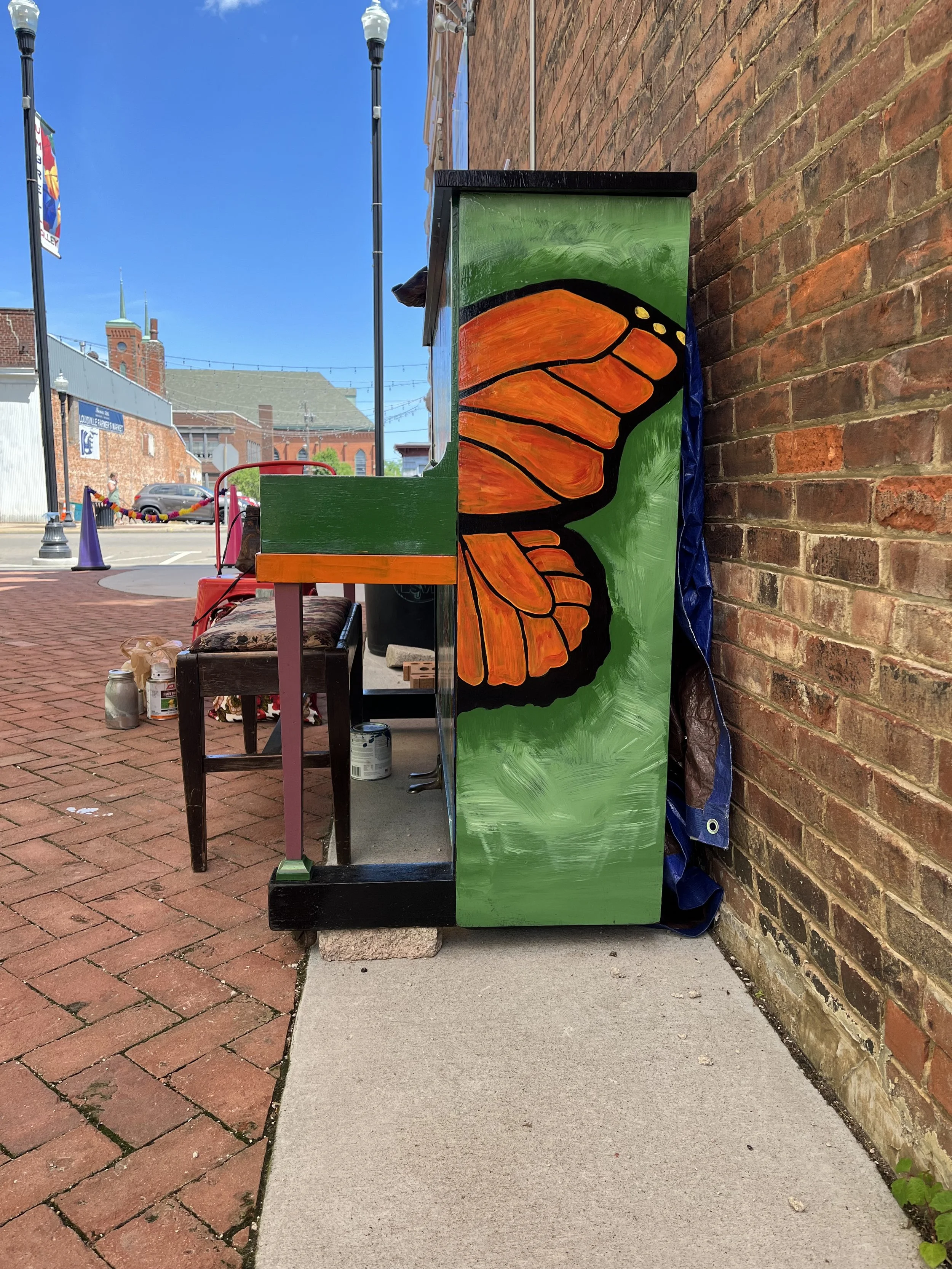 A painted wooden structure featuring an orange butterfly wing design, situated outdoors on a brick sidewalk next to a brick wall with street lights and buildings in the background.