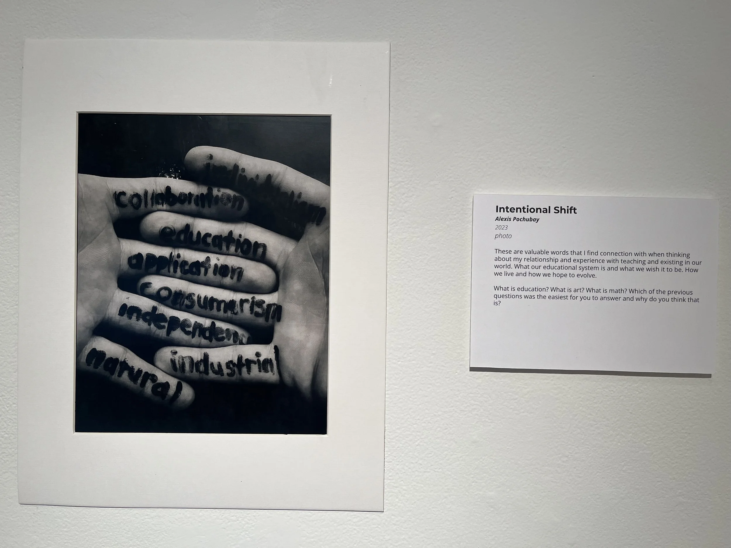 Black and white photograph of a person holding a finger close to the camera, with words written on the finger including 'collaboration', 'education', 'application', and 'industry'.