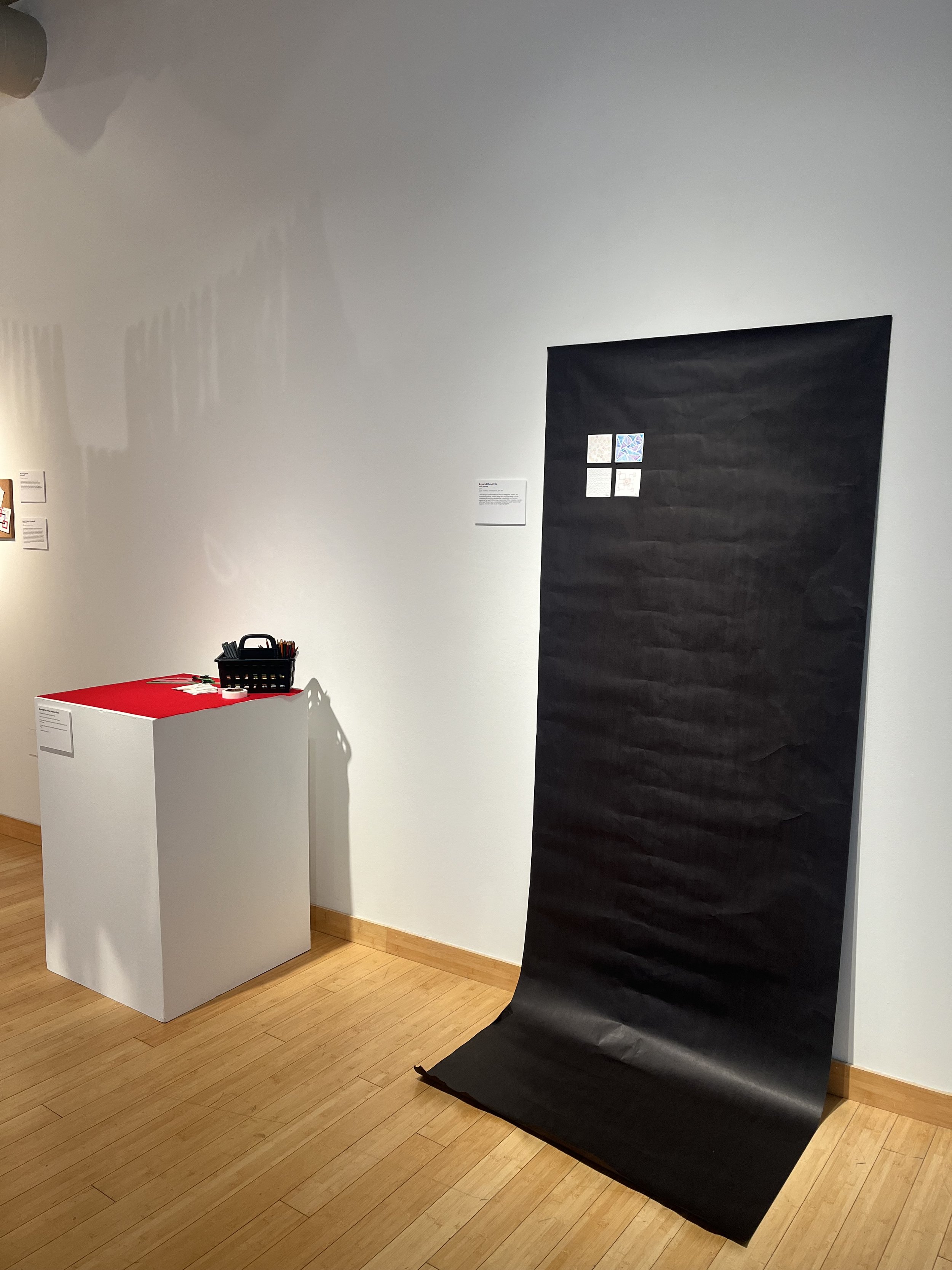 An art exhibit featuring a black textured paper backdrop, a white pedestal with a red cloth on top, and art supplies in a black basket, displayed in a gallery with wooden flooring and white walls.