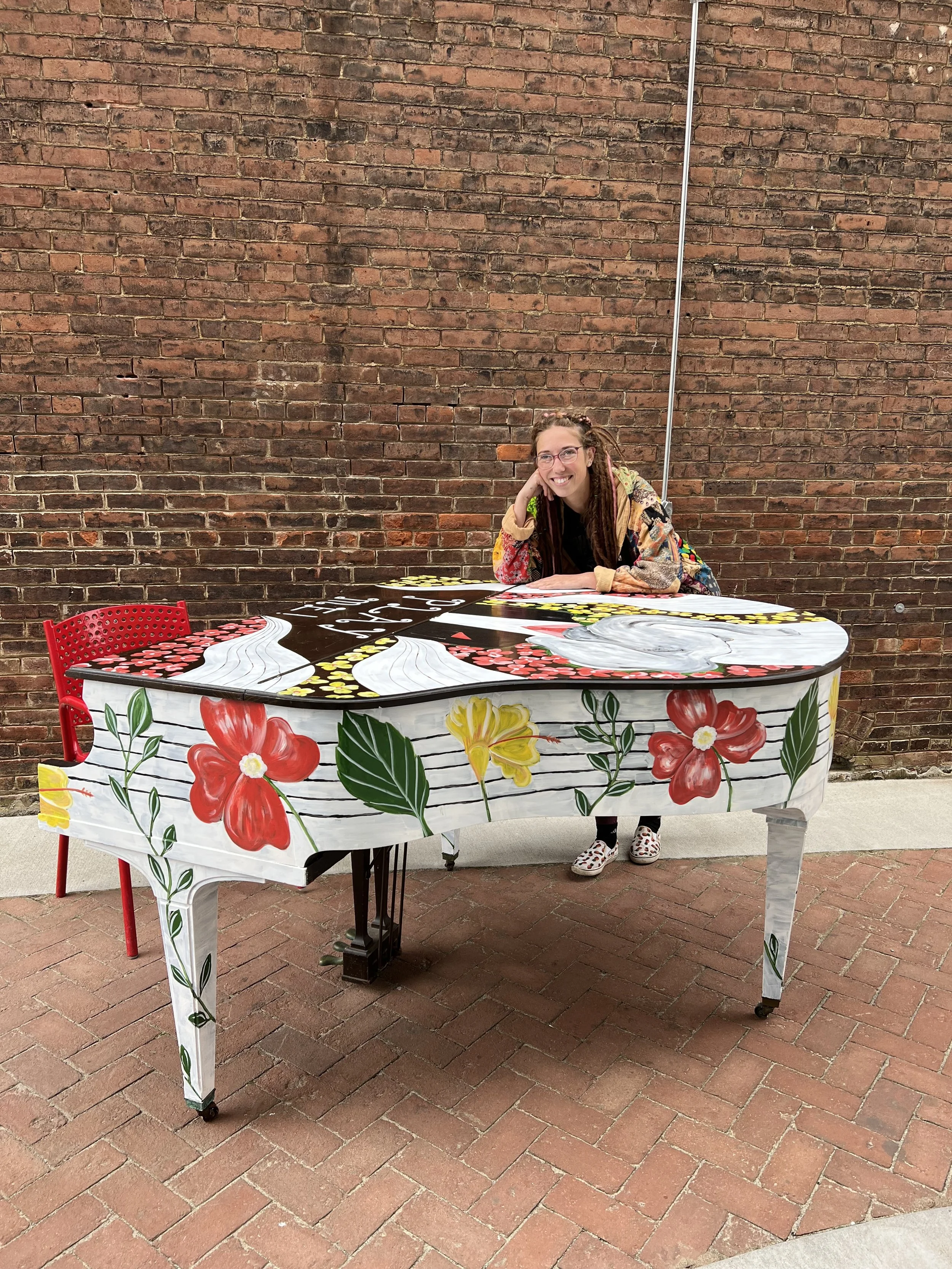 A woman with glasses and dreadlocks, wearing a colorful patchwork jacket and polka dot shoes, is sitting and leaning on a decorated digital piano with floral and fruit designs. She is smiling and resting her head on her hand, in front of a brick wall