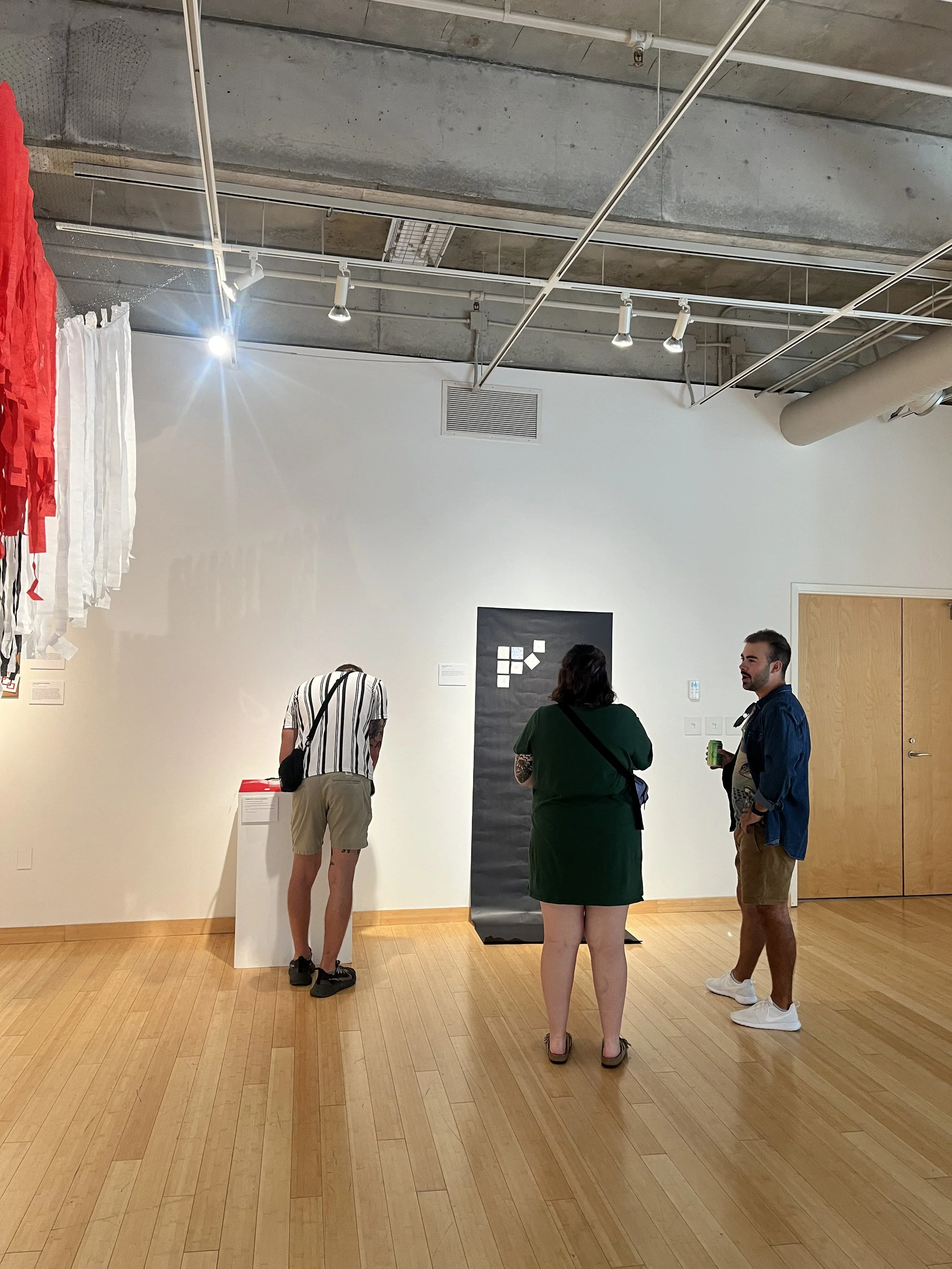 Four people are standing and talking in an art gallery with wooden floors and white walls. One person, wearing a striped shirt, is looking at artwork on the wall. A woman in a green dress and two men, one in a dark shirt and khaki shorts and another 