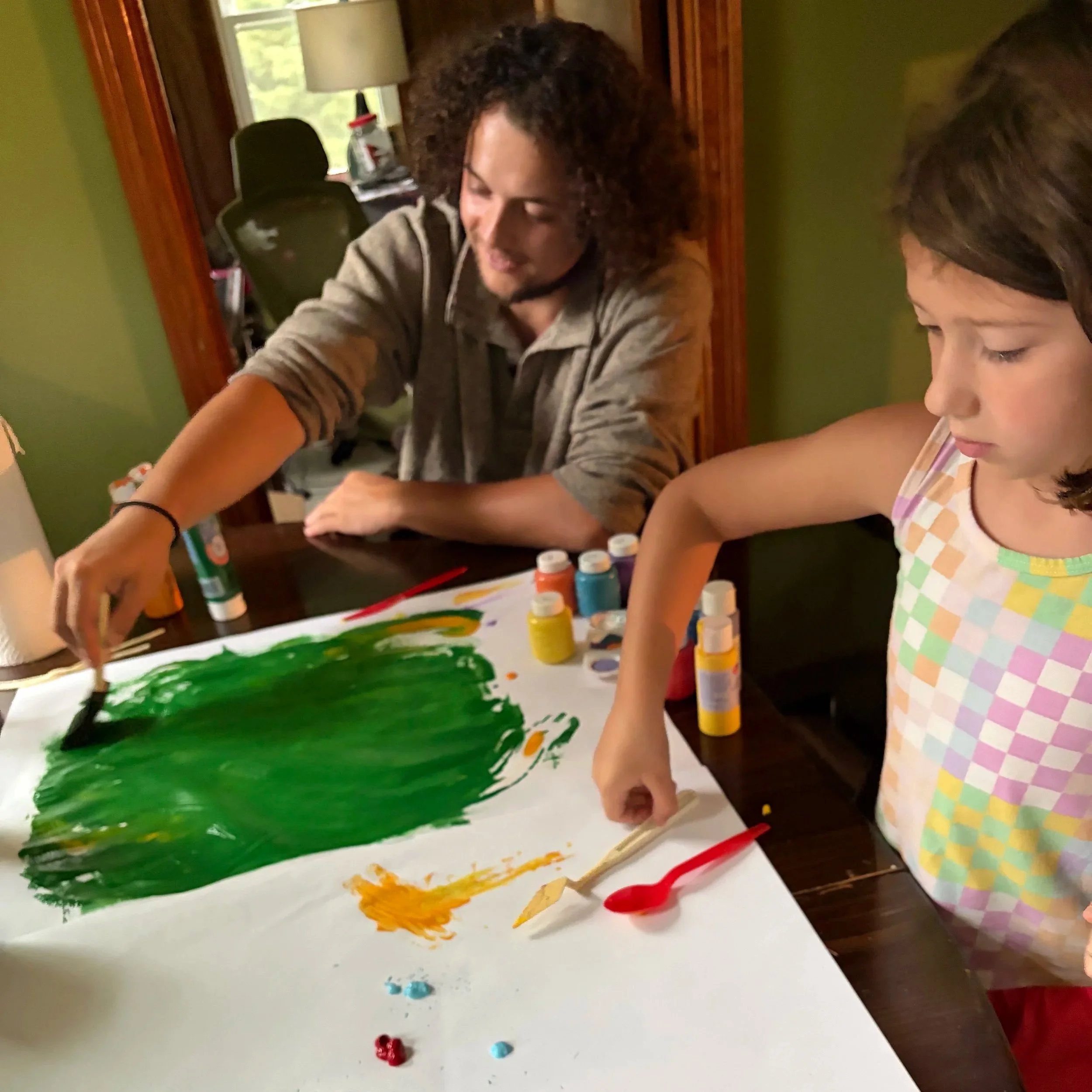 A young girl in a sleeveless top is painting on a large sheet of paper with green, yellow, and orange acrylic paints. An adult male with curly hair and a beard, wearing a hoodie, is assisting her. Acrylic paint bottles and paintbrushes are on the table. The scene takes place in a room with a green wall and a window in the background, and a mirror reflecting part of the room.