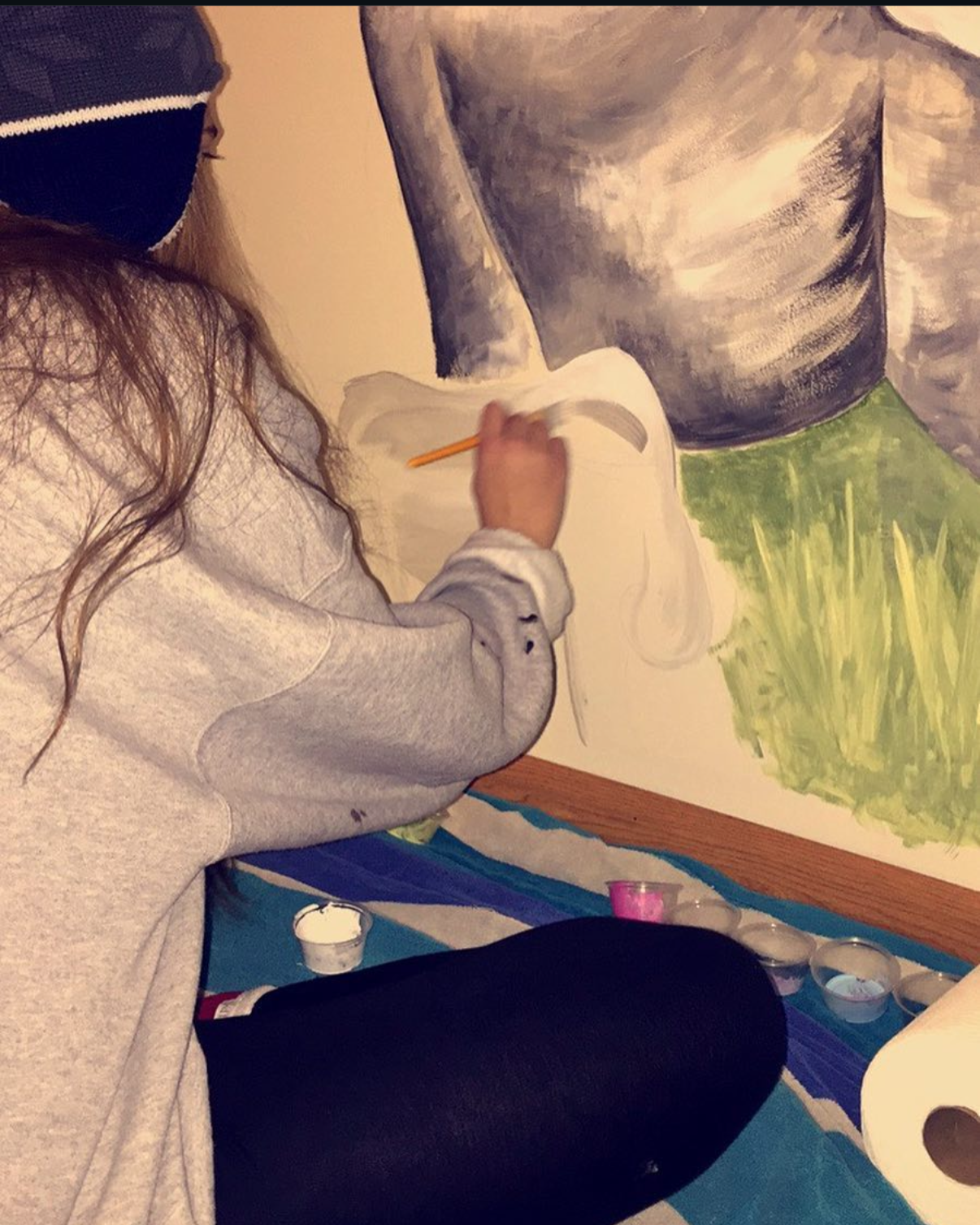 A person with long hair, wearing a gray sweatshirt and a beanie, is sitting on the floor and painting a large mural of a person’s torso and grass on the wall.