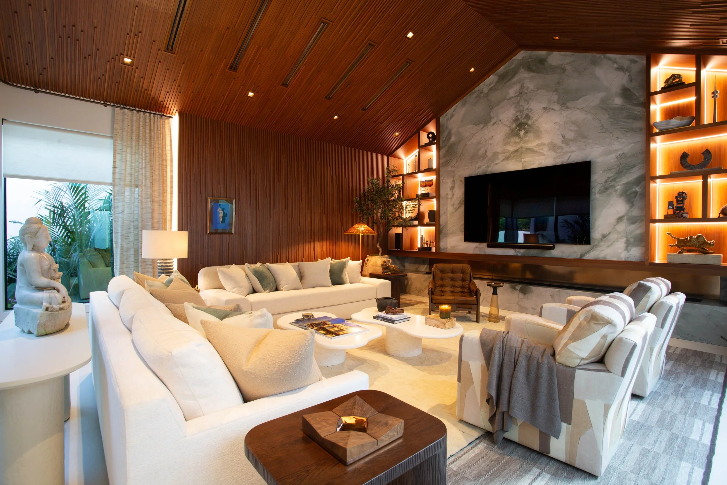 Interior of a modern living room with white sofas, a stone fireplace, wooden ceiling, built-in shelves with decor, large window with curtains, and various decorative items.