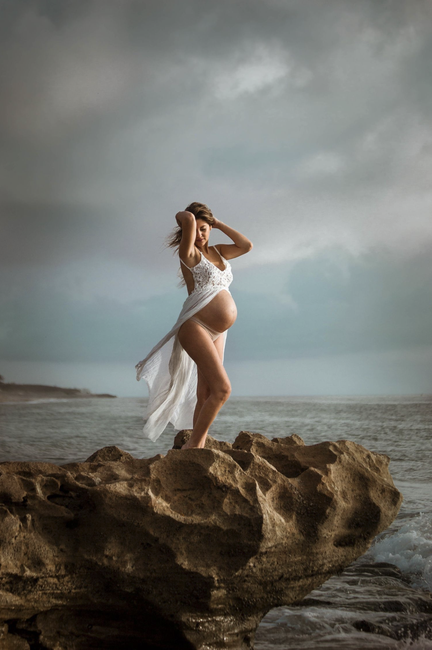 A pregnant woman standing on a large rock by the water at the beach, wearing a white lace dress. The sky is cloudy with a mix of dark and light clouds.