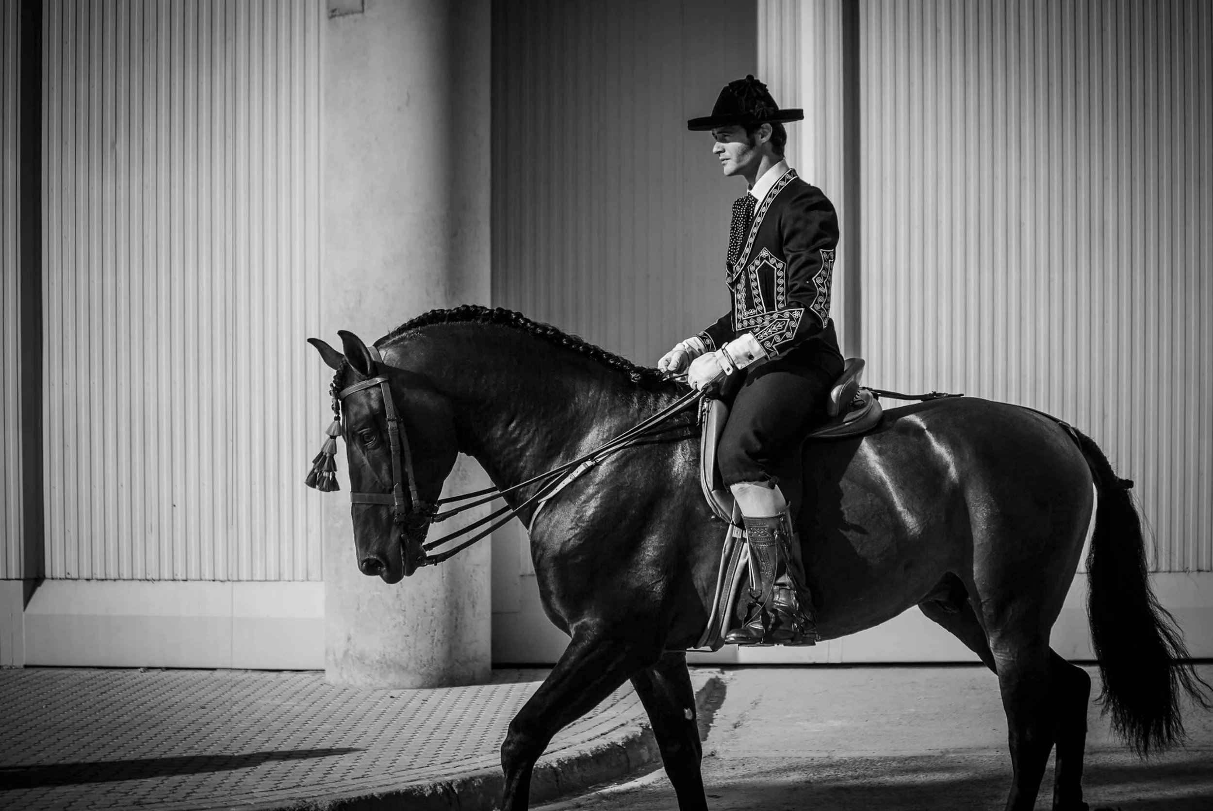 A person riding a horse inside an indoor riding arena, dressed in traditional equestrian attire with a wide-brimmed hat and ornate jacket.