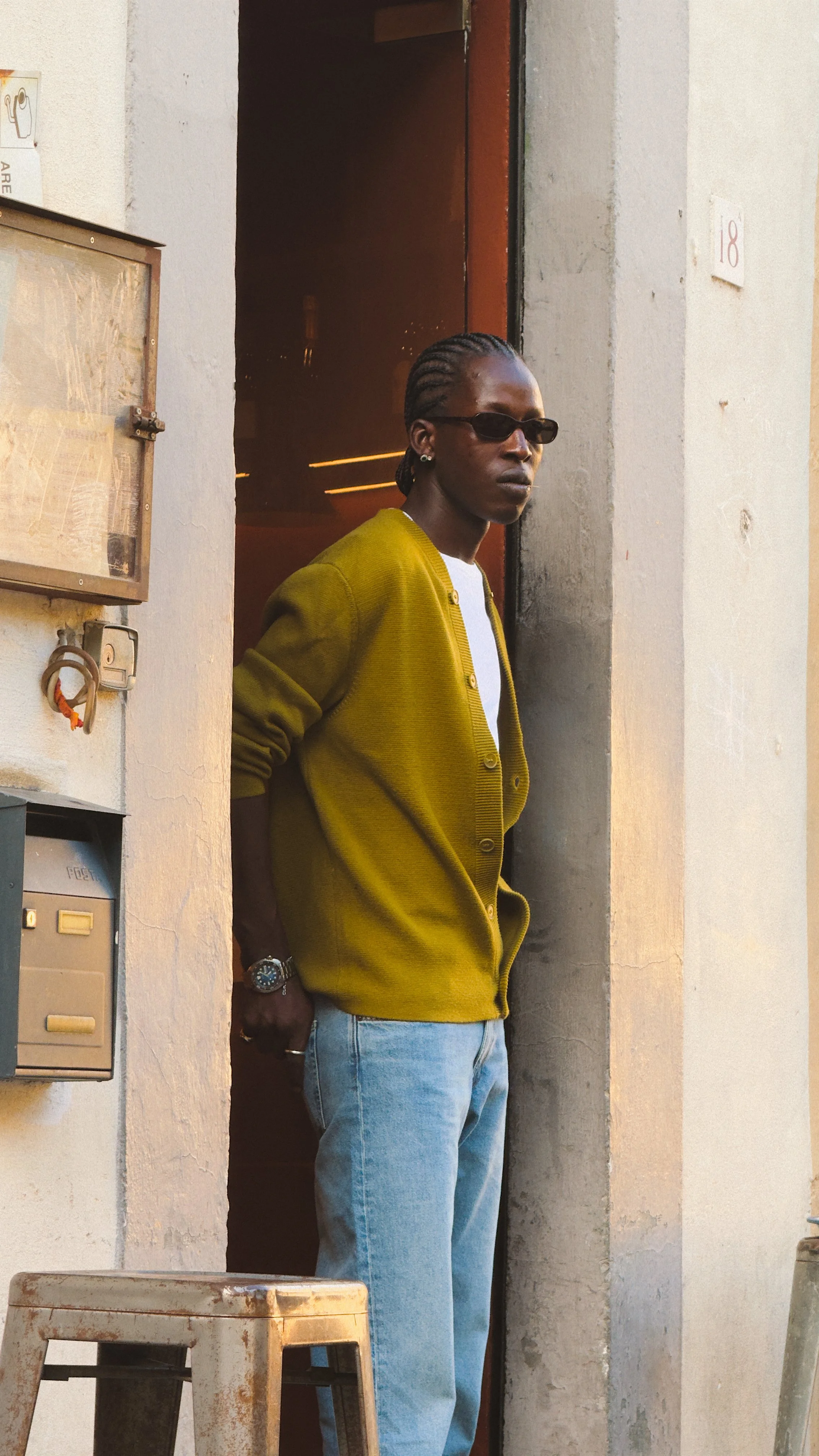 A person with braided hair wearing sunglasses, a green cardigan, and jeans standing in a doorway outside a building.