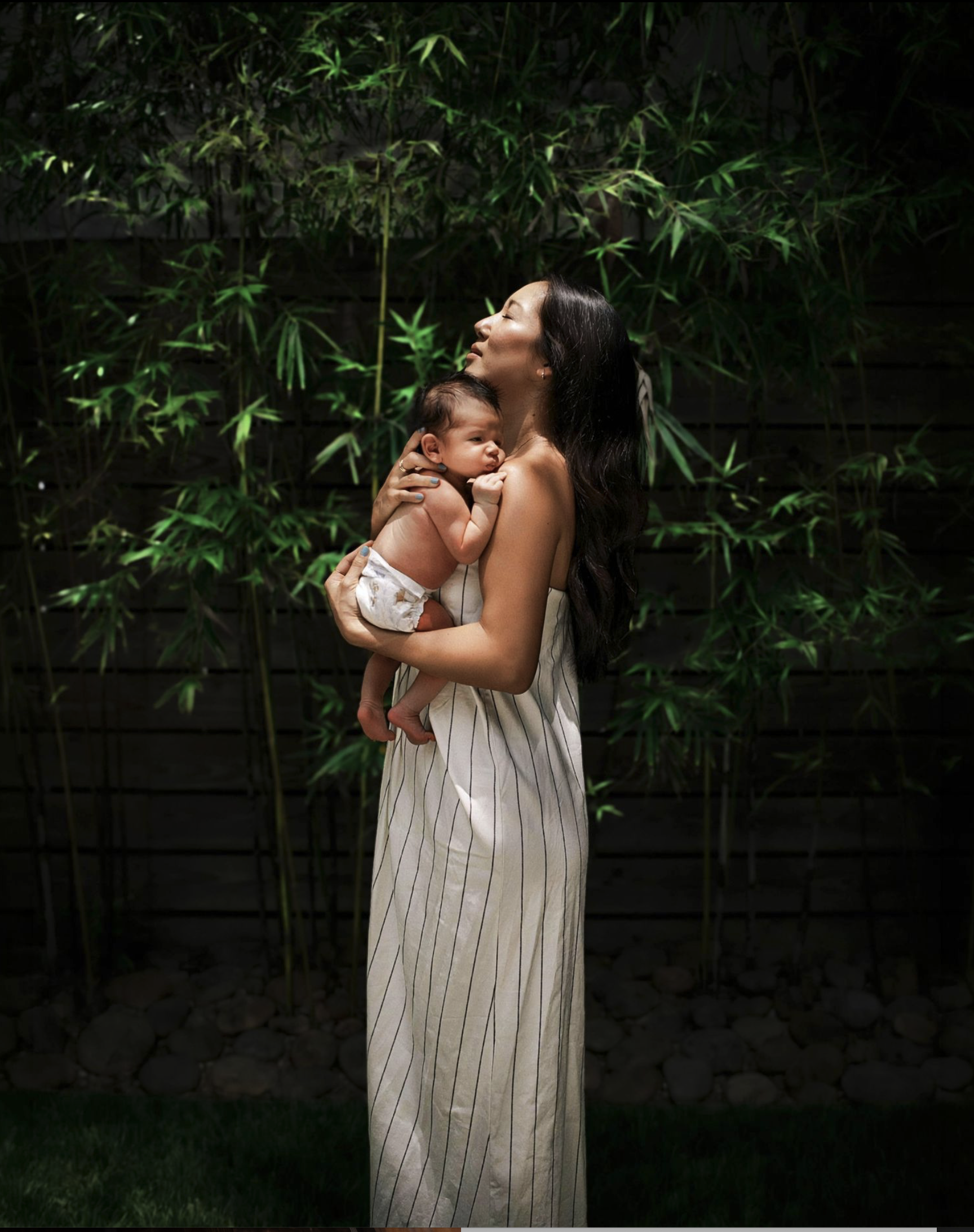 A woman with long dark hair holding a baby with short dark hair, both with their eyes closed, standing in front of a green leafy background.