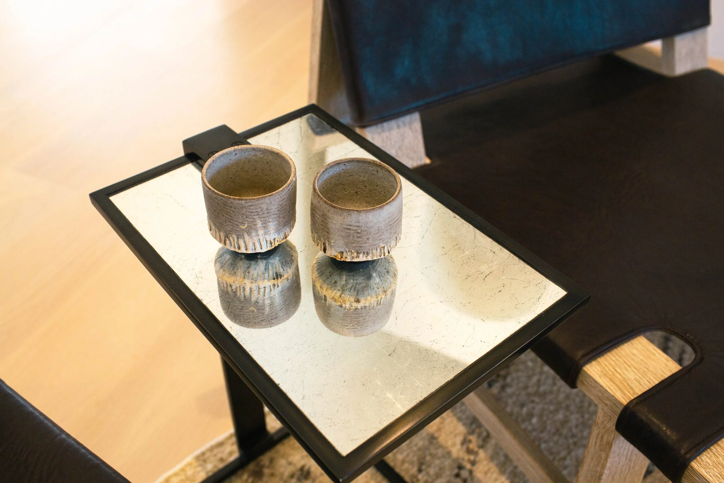 Two beige ceramic cups with textured bottoms placed on a reflective surface, on a black metal table in an indoor setting.